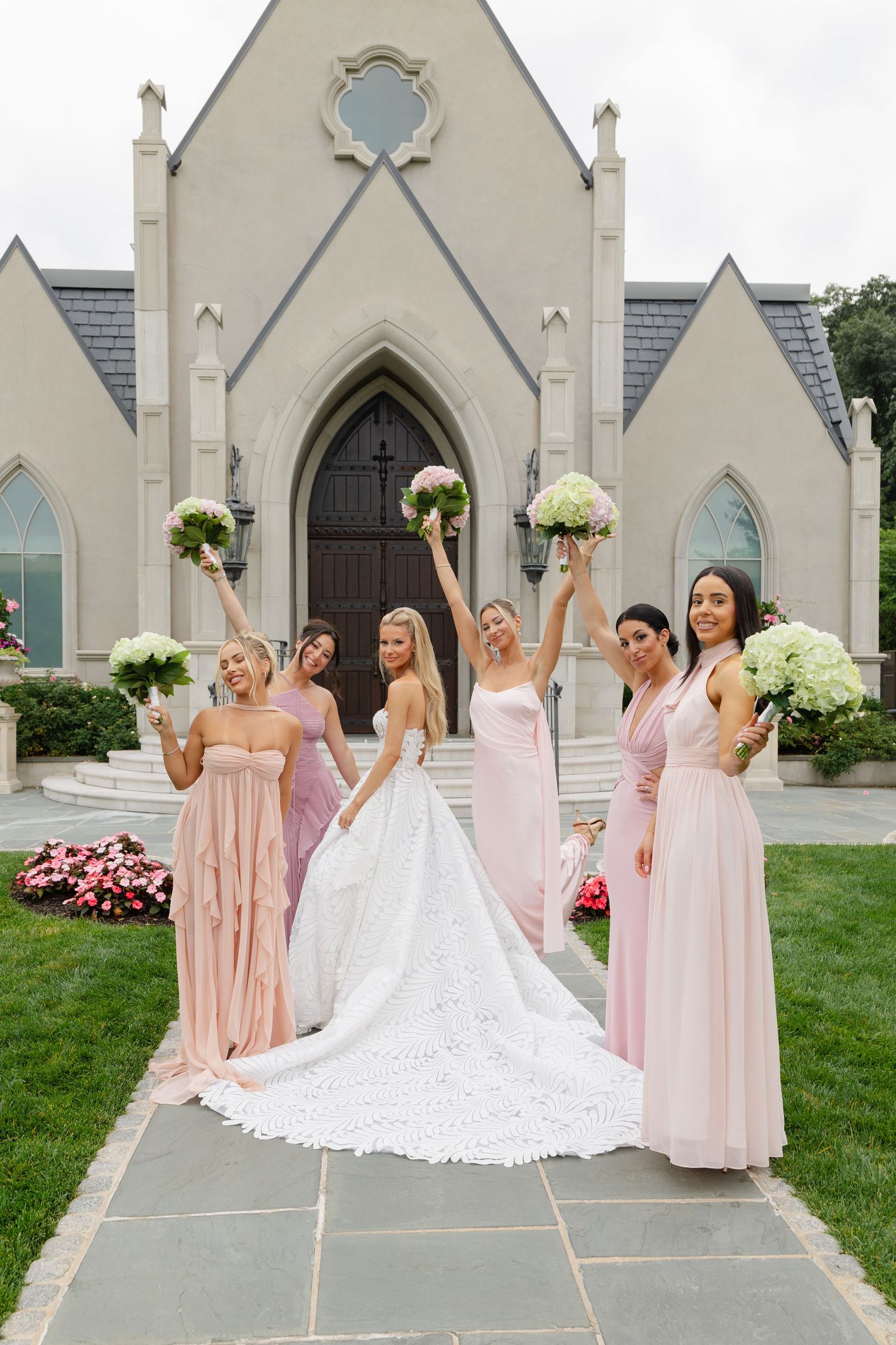 Bride in white gown with five bridesmaids in pink dresses celebrating outside a stone chapel, holding bouquets.