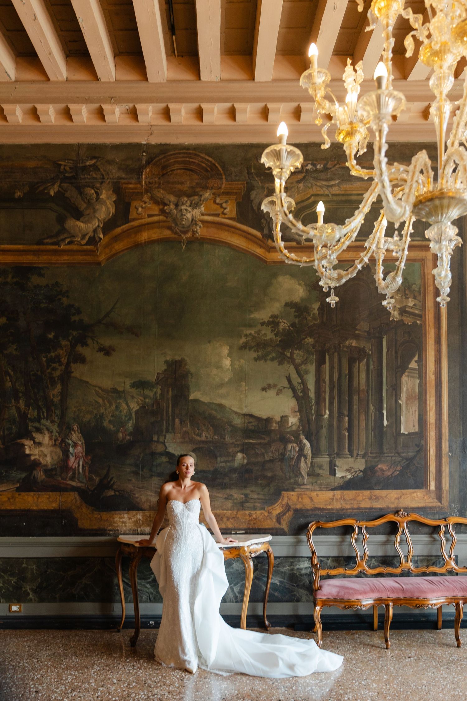 Elegant white wedding dress photographed in ornate room with vintage mural and crystal chandelier in luxury European villa.