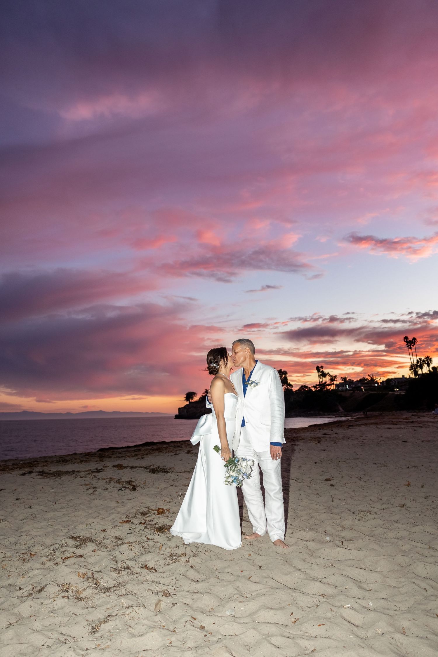 Wedding couple silhouetted against a dramatic purple and pink sunset on a sandy beach.