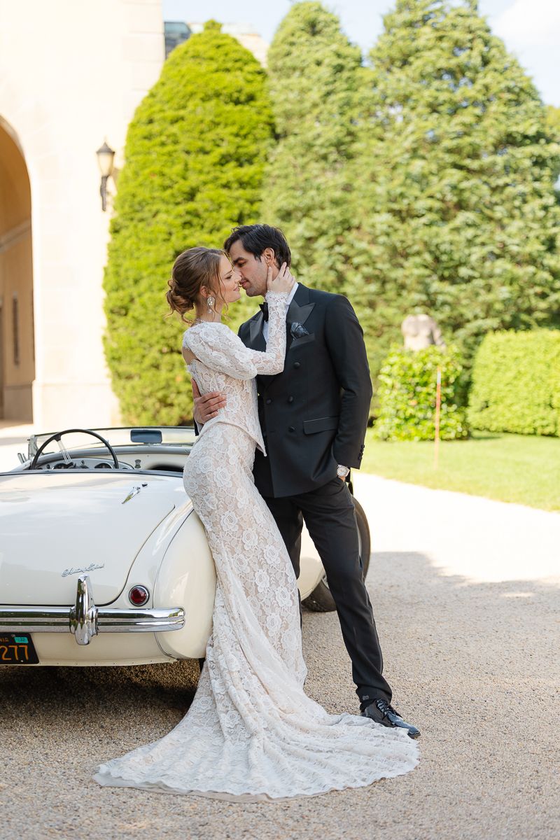 A couple poses romantically next to a vintage white car on their wedding day.