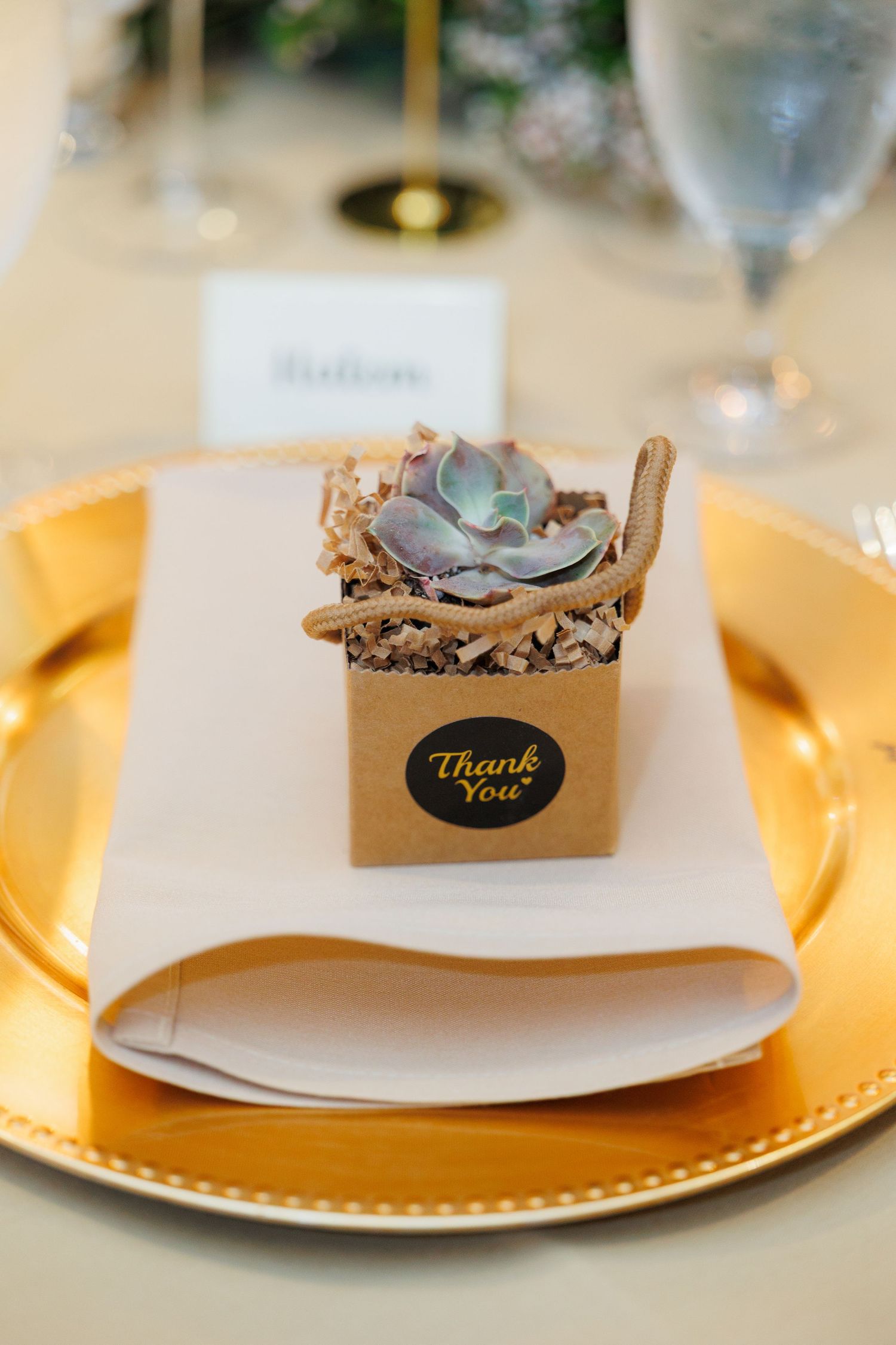 A succulent wedding favor in a brown box with 'Thank You' sticker sits on a gold charger plate with white napkin.