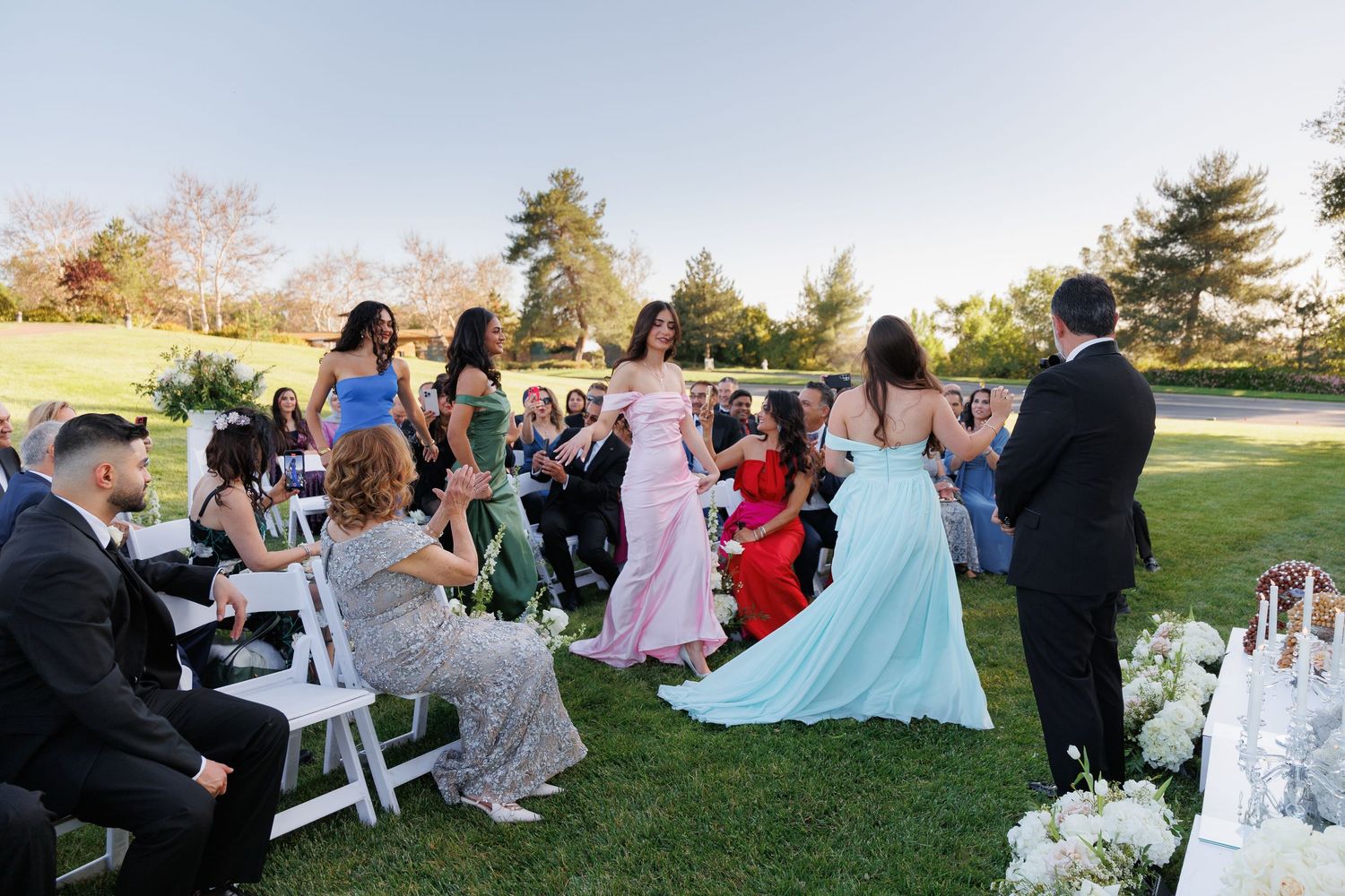 Wedding guests sit in white chairs on a lawn during an outdoor ceremony at sunset.