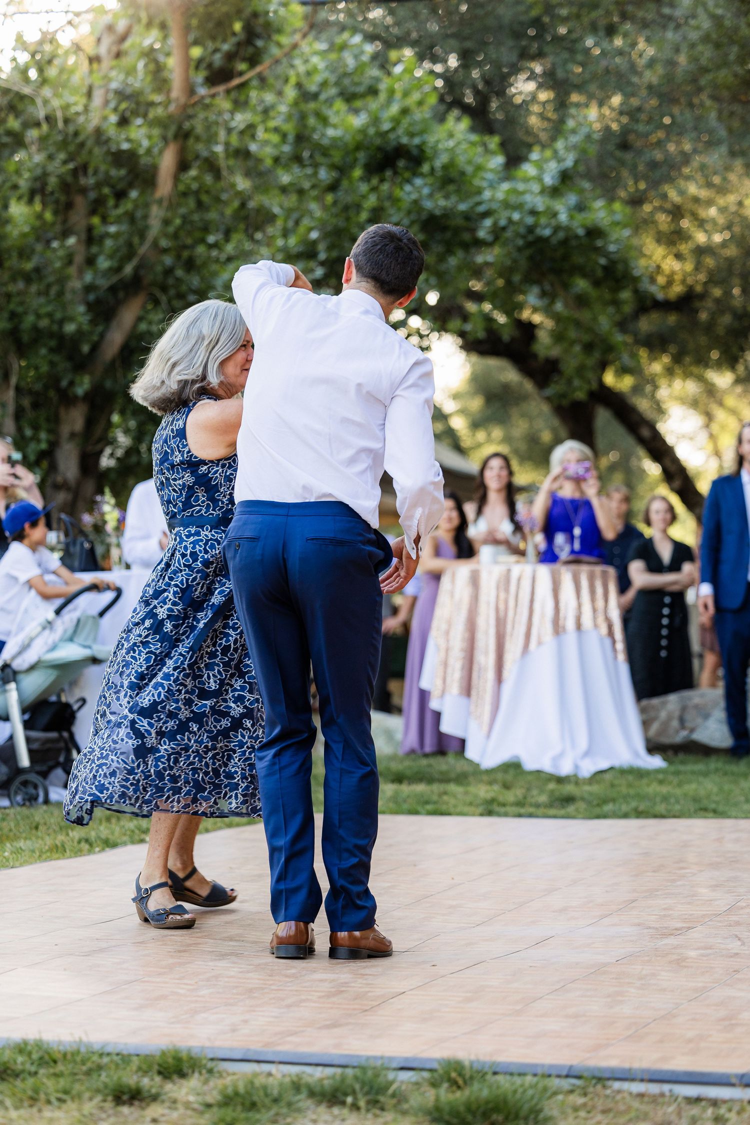 Guests dance on an outdoor wooden dance floor at a garden wedding reception with trees in the background.