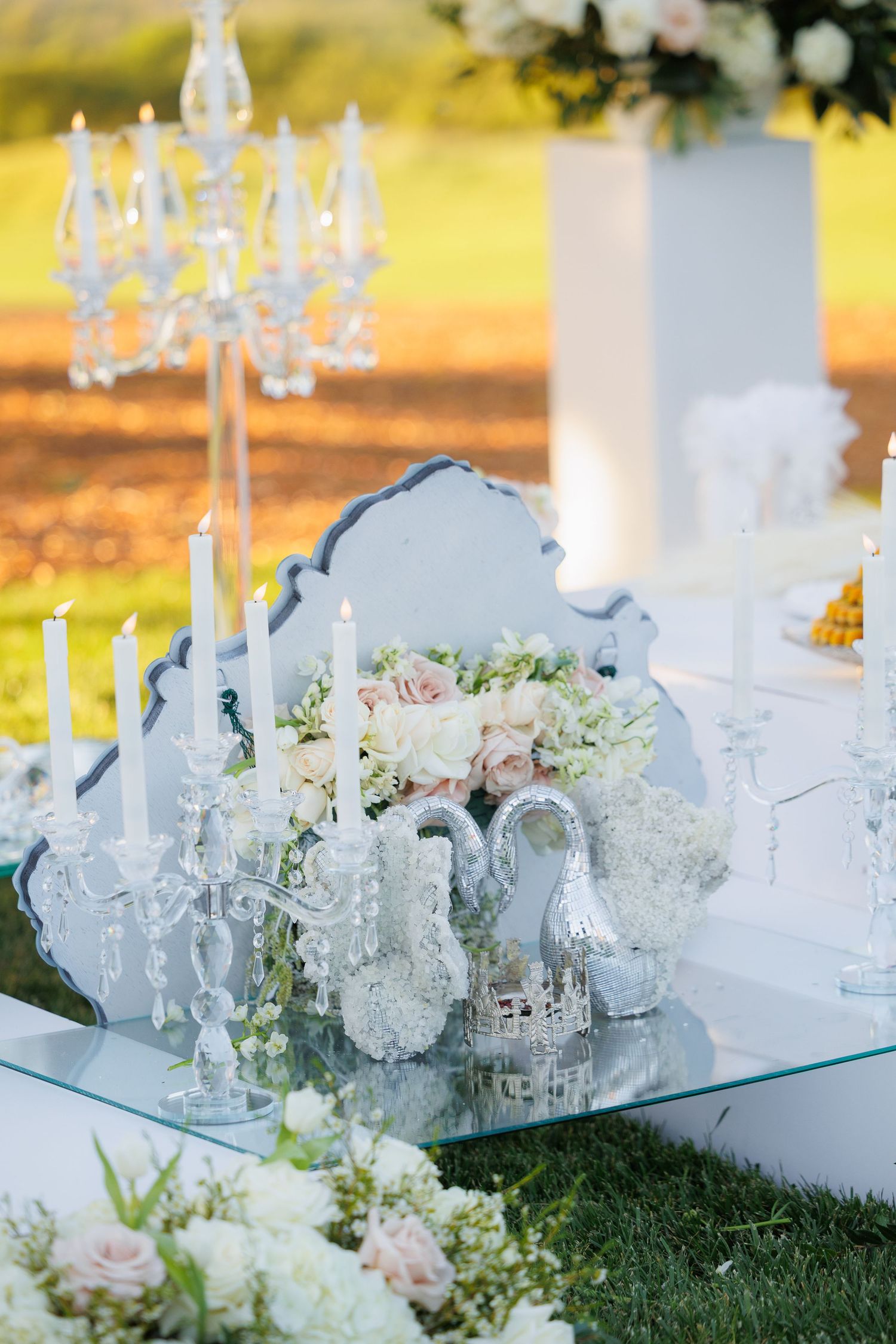 Elegant outdoor wedding decor with crystal candelabras, white flowers, and mirrored accents on a glass table.