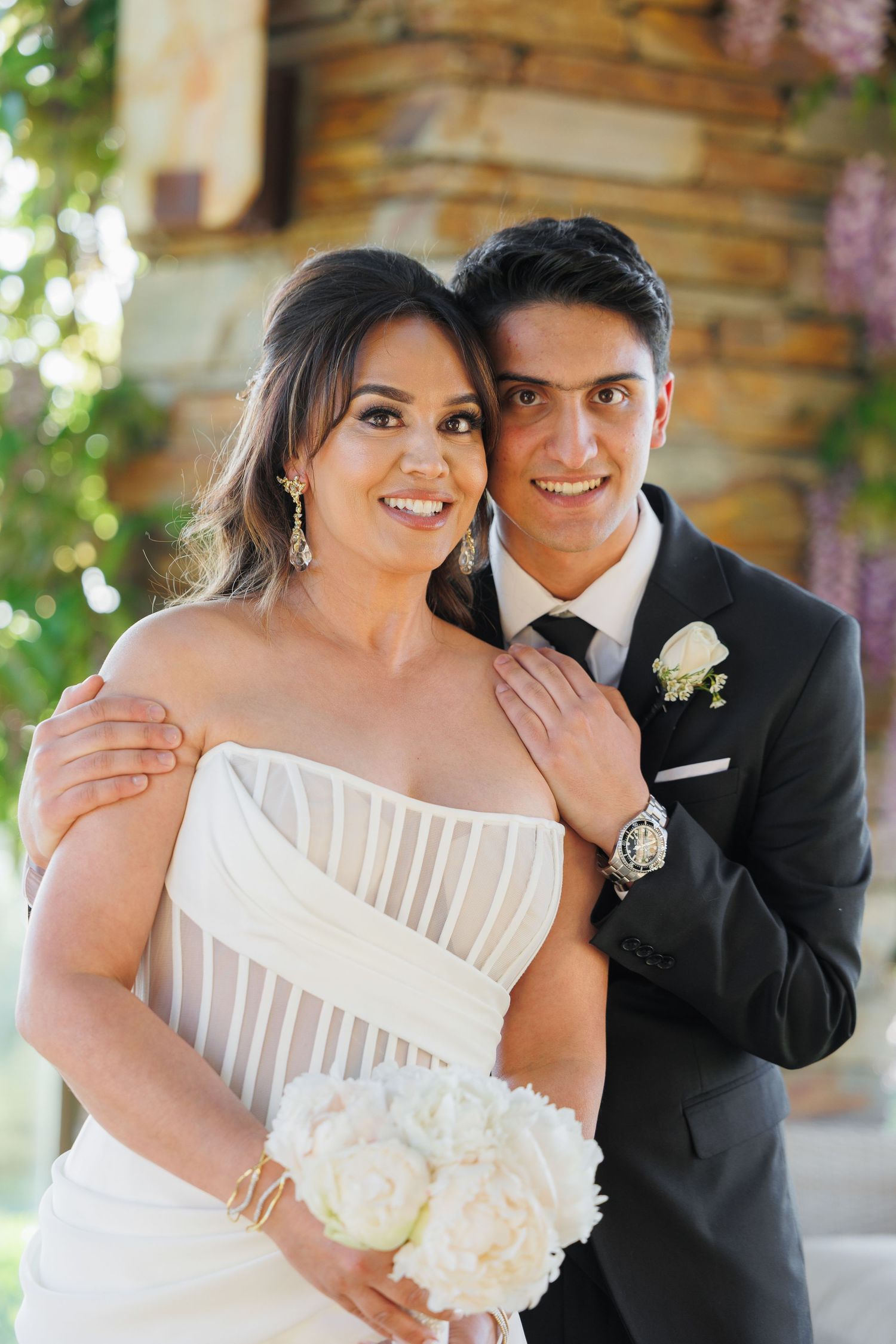 Romantic wedding couple in formal attire embracing and smiling outdoors near rustic wooden structure.