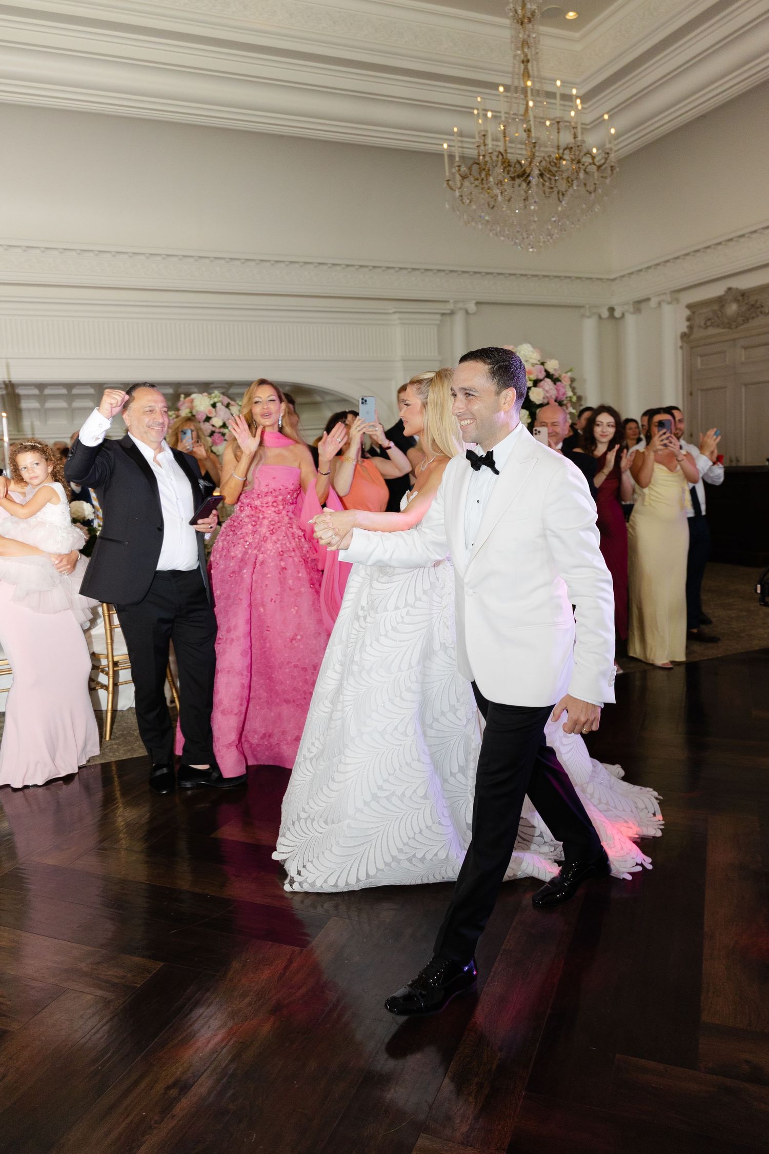 Couples dancing in formal attire at an elegant ballroom event with chandeliers and hardwood floors.