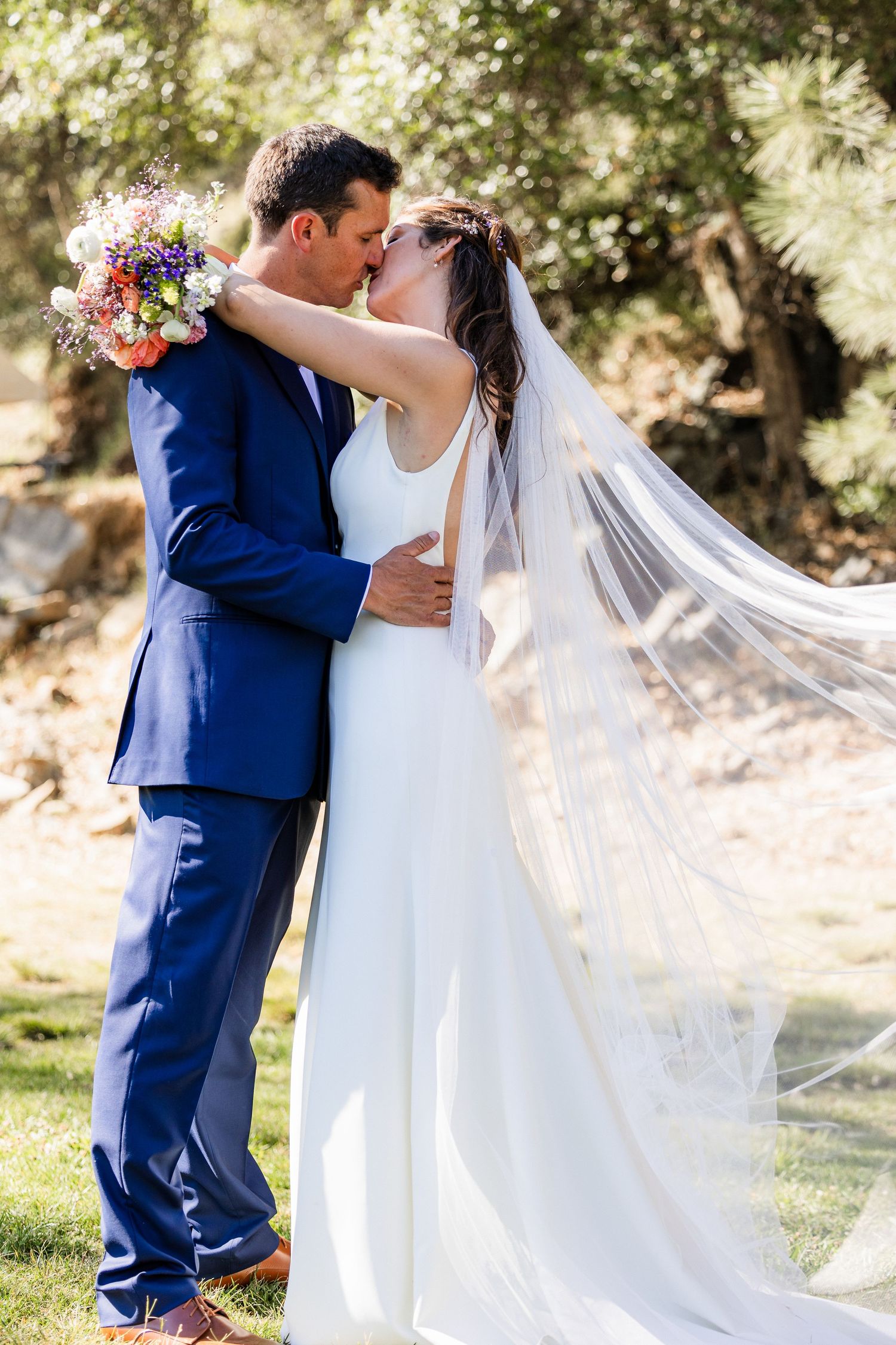 A romantic wedding couple embraces outdoors in a garden setting with flowing white dress and navy suit.