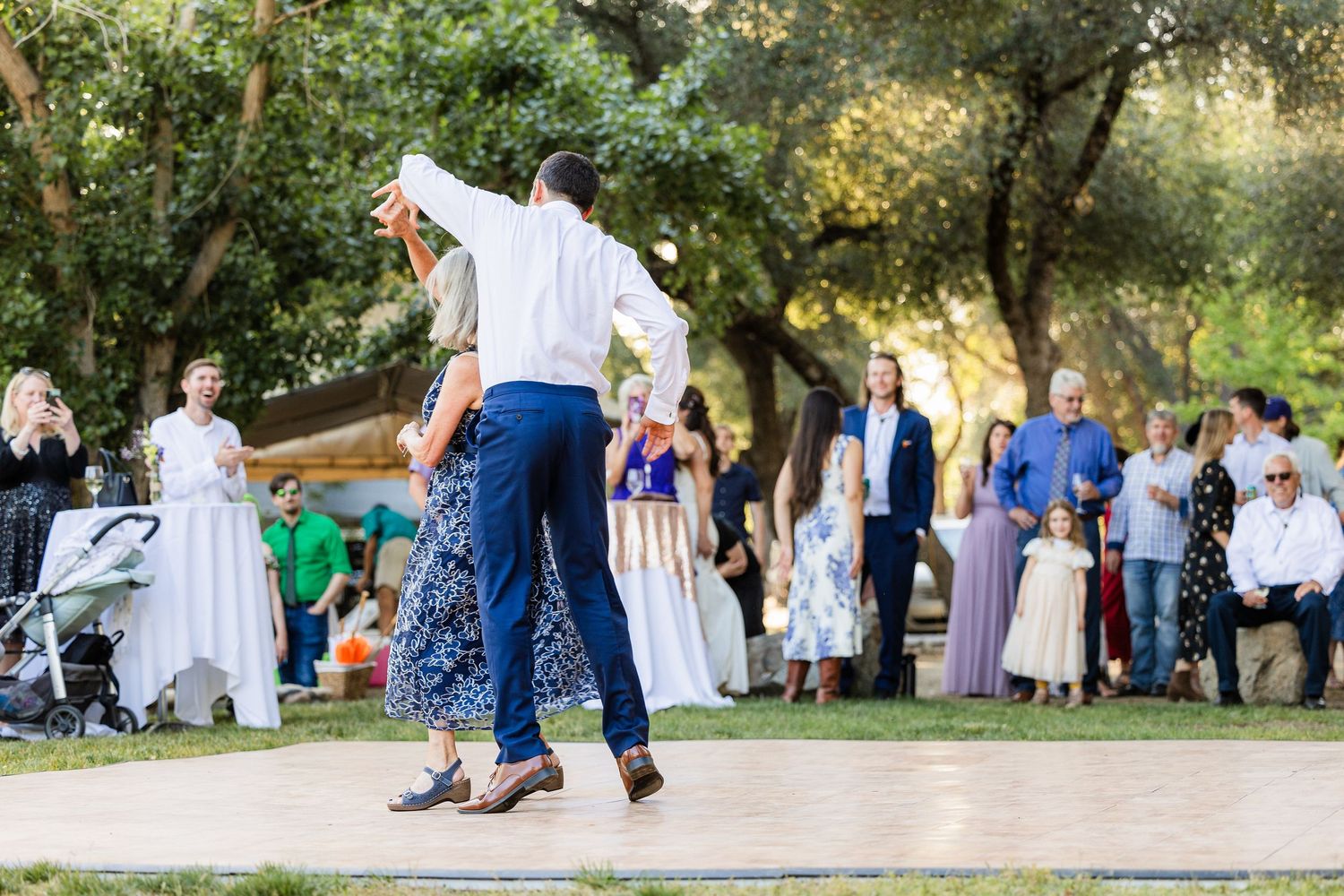A group of people dancing in a line outdoors on a gravel path surrounded by green trees and natural scenery.