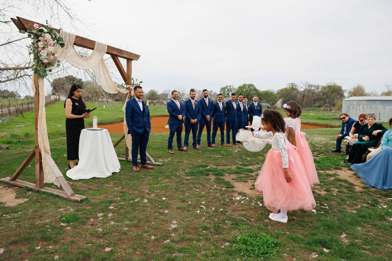 Wedding guests gather outdoors on a grassy hill under wooden archways for a rustic outdoor ceremony.