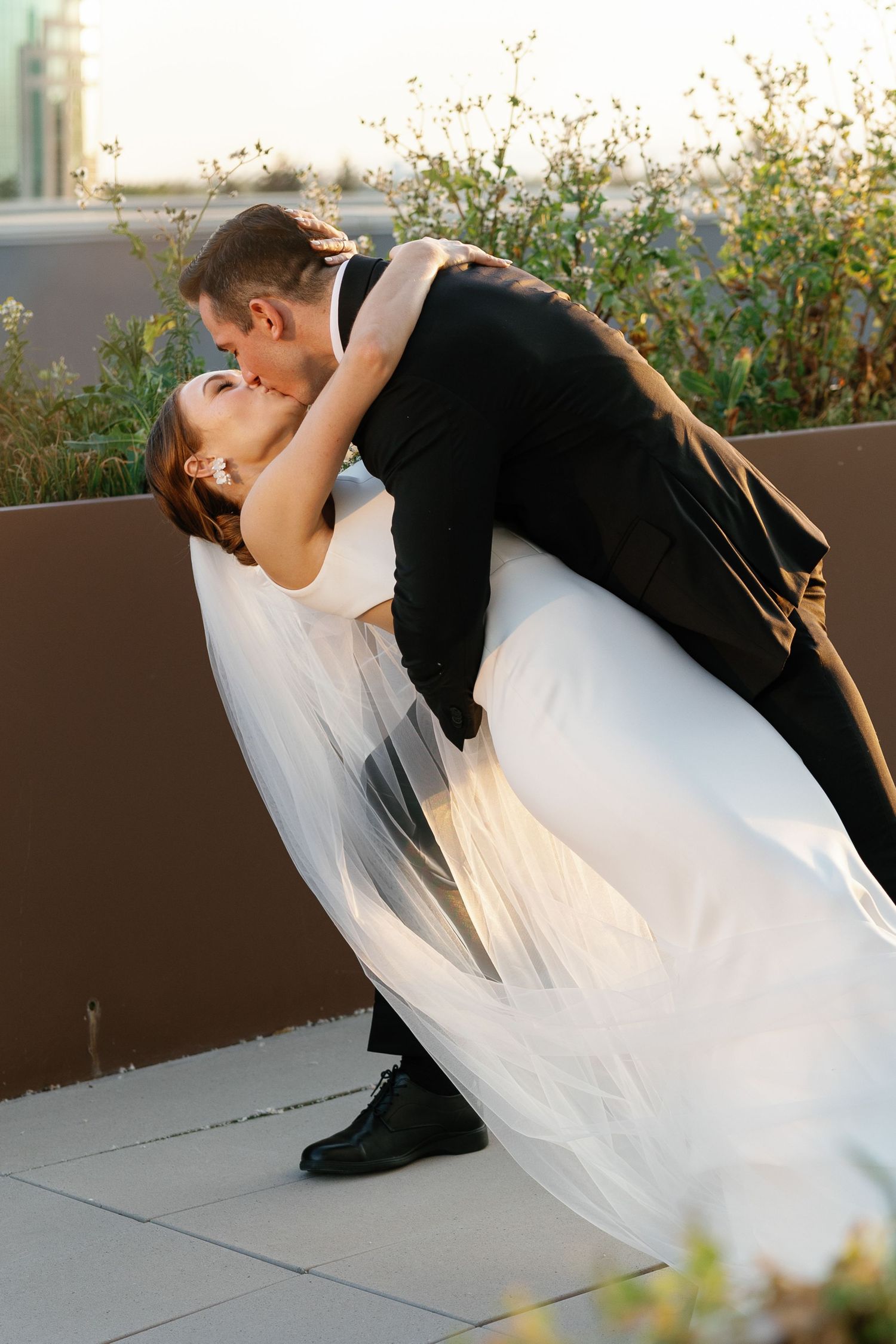 A romantic wedding couple dips during their dance on a rooftop terrace at sunset with flowing white veil.