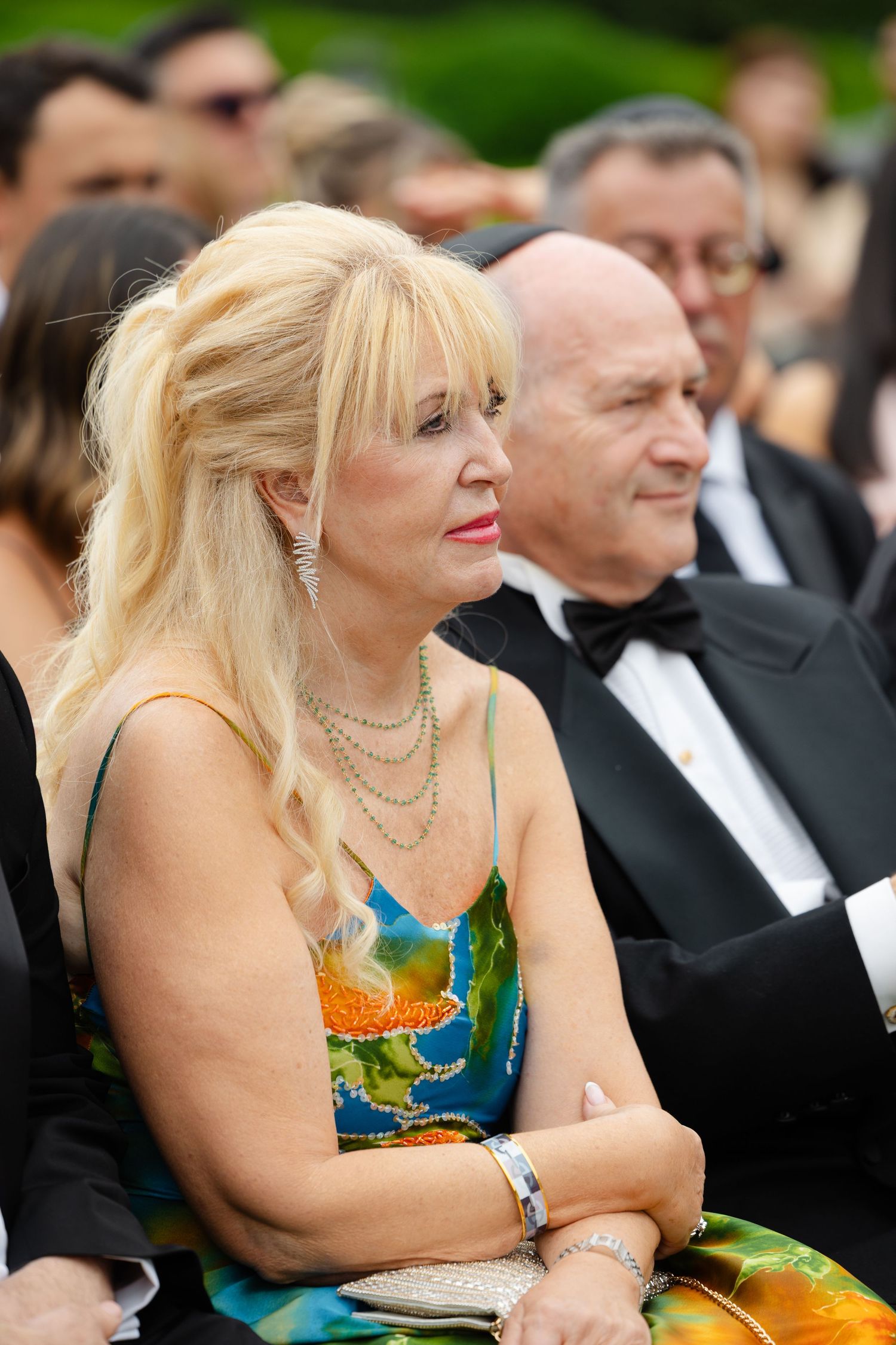 Attendee in colorful blue and green dress with pearl necklaces watches intently at formal outdoor event.