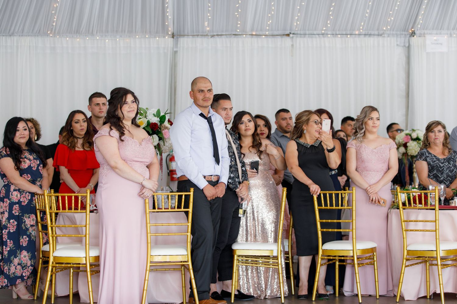 Wedding guests stand during ceremony in a white tent venue with gold chiavari chairs.