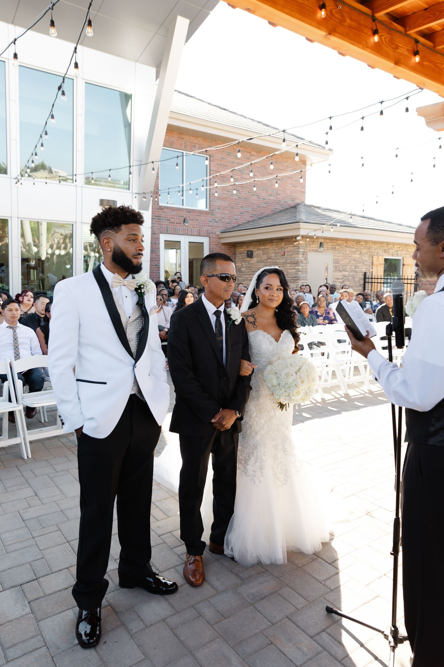 Wedding party guests mingle and celebrate on an elegant outdoor patio with string lights and glass walls.