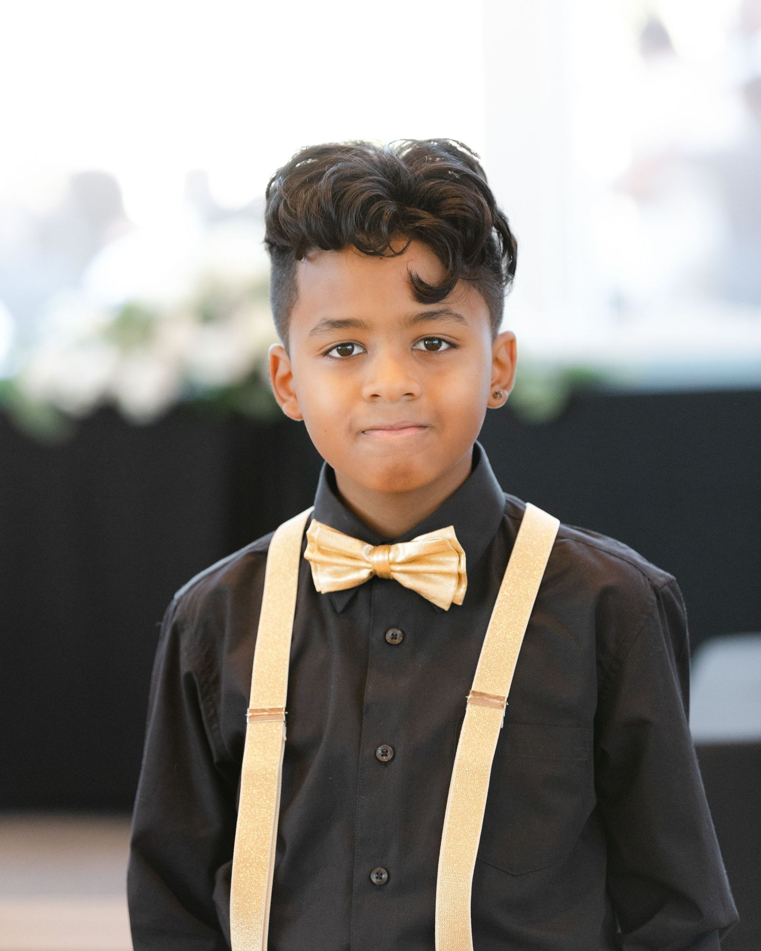 Young child wearing a formal black suit with beige suspenders and bow tie smiles warmly at the camera.