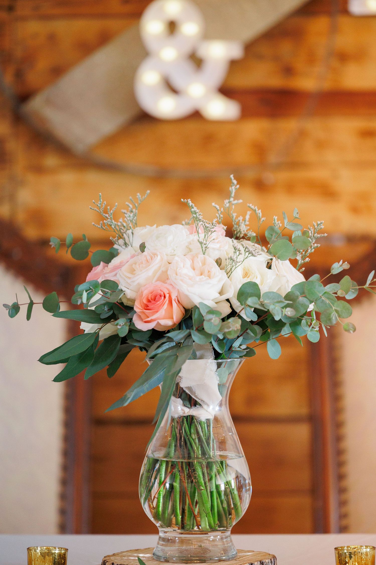 Pink and white roses with eucalyptus arranged in a glass vase create an elegant wedding centerpiece on a wooden table.