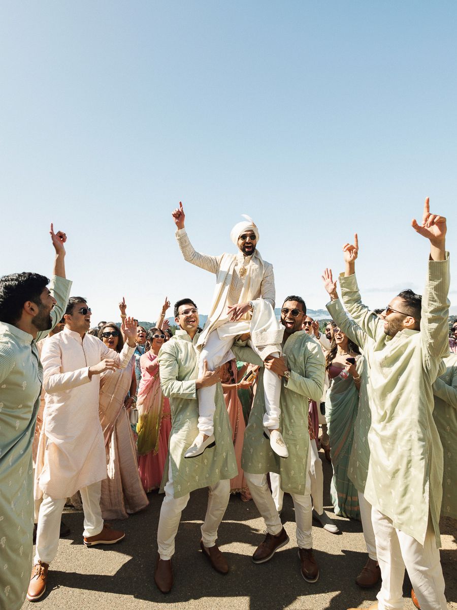 Groomsmen in light colored traditional outfits celebrating with raised arms against blue sky.