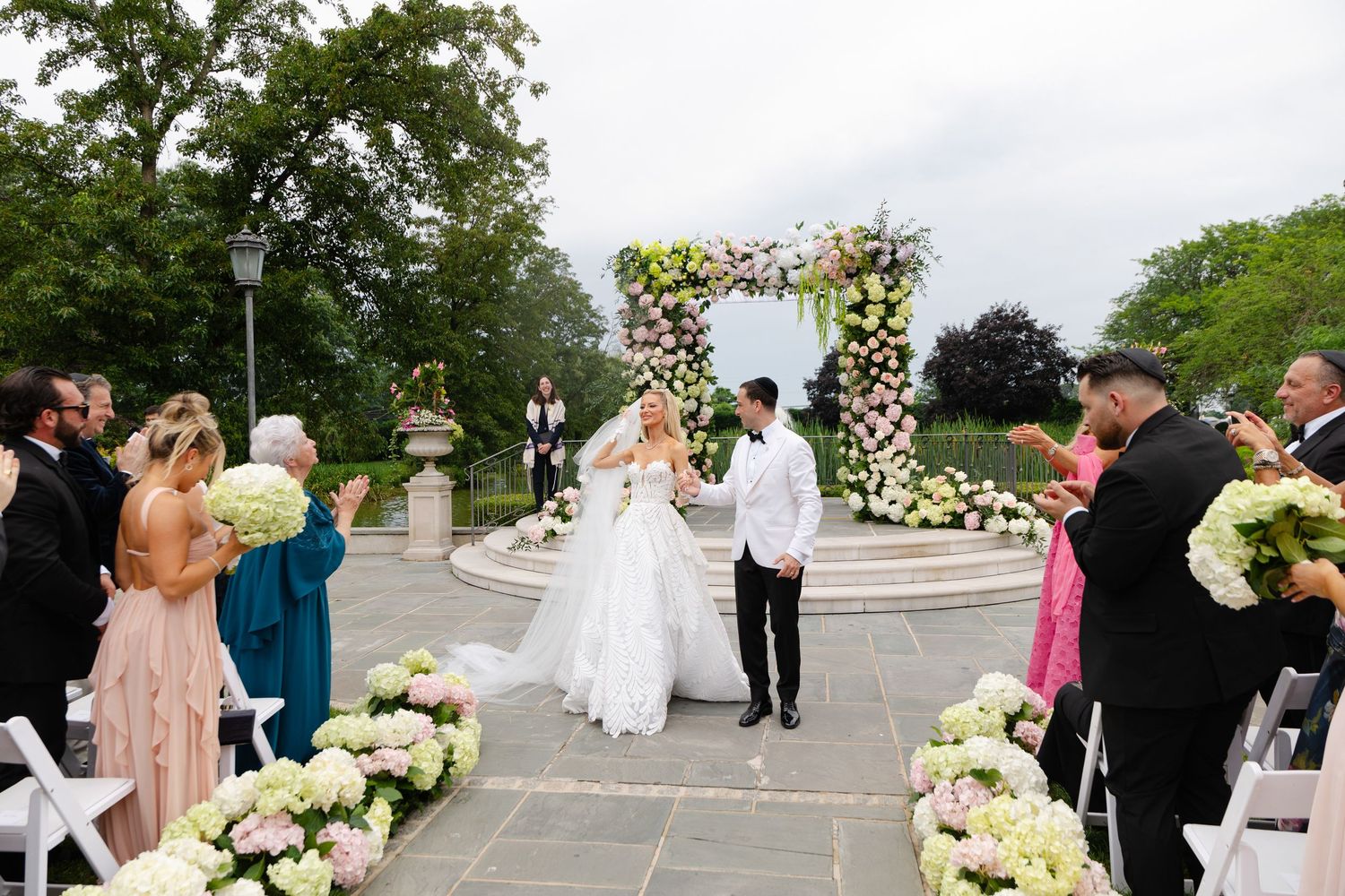 Bride and groom walk down the aisle at an outdoor wedding ceremony with floral arch and hydrangea arrangements.
