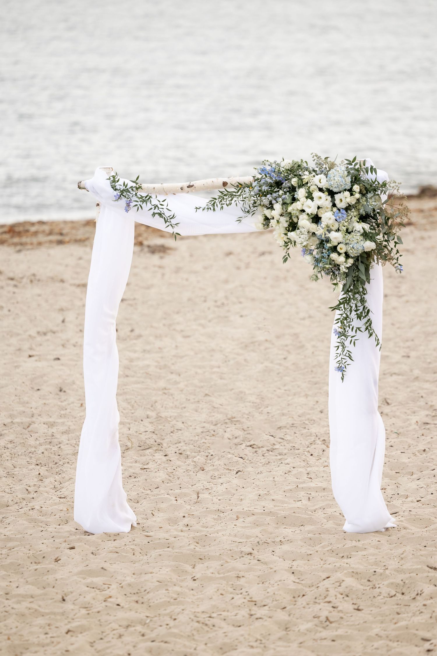 White fabric drapes decorated with delicate floral arrangements create an elegant beach wedding arch on sandy shore.