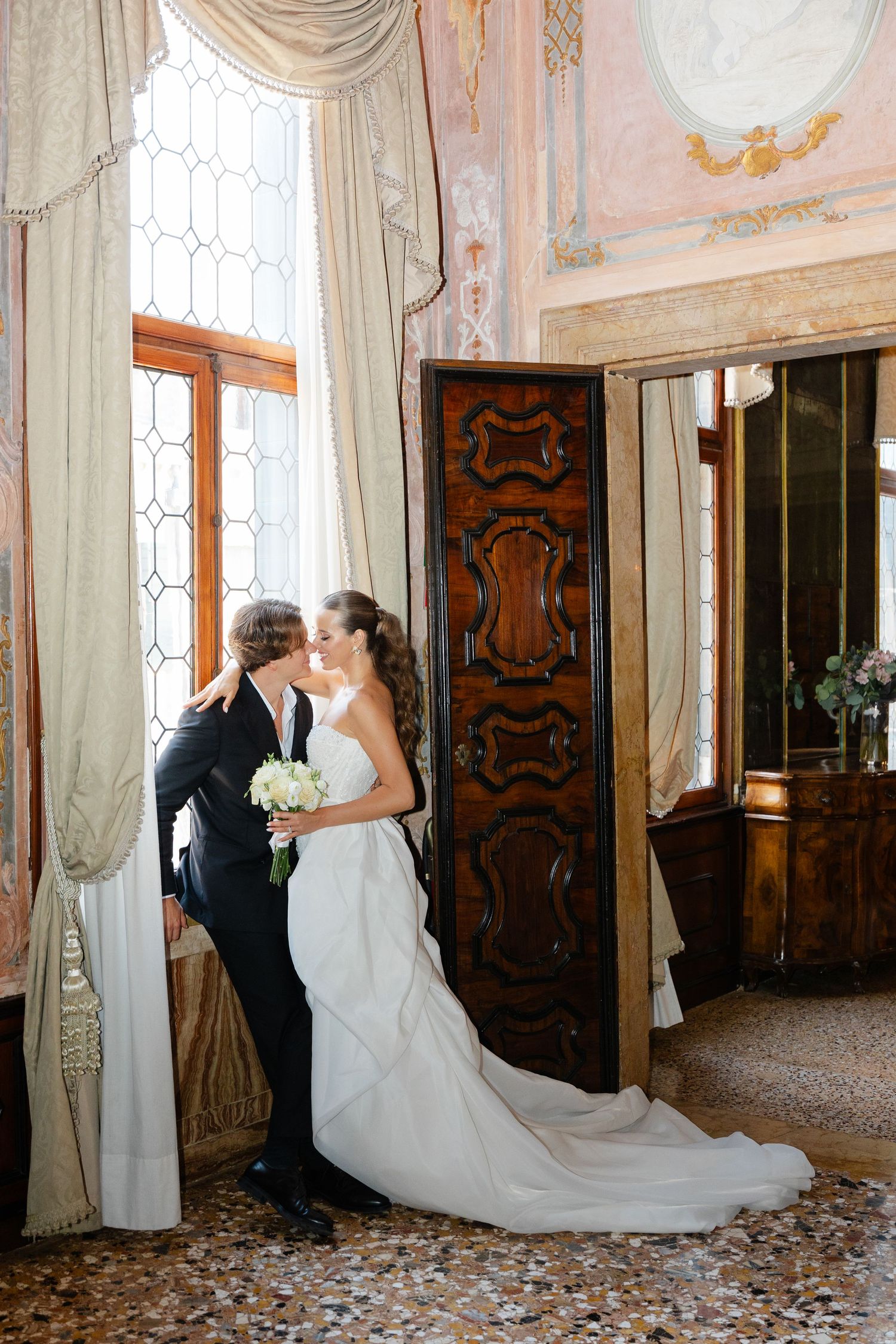 Elegant couple embraces in formal attire beside ornate wooden door and flowing curtains in historic interior setting.