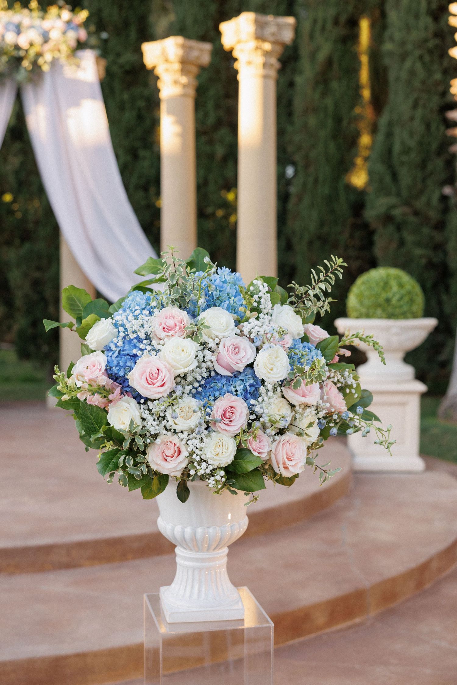 An elegant floral arrangement of pink roses and blue hydrangeas in a white urn against classical columns.