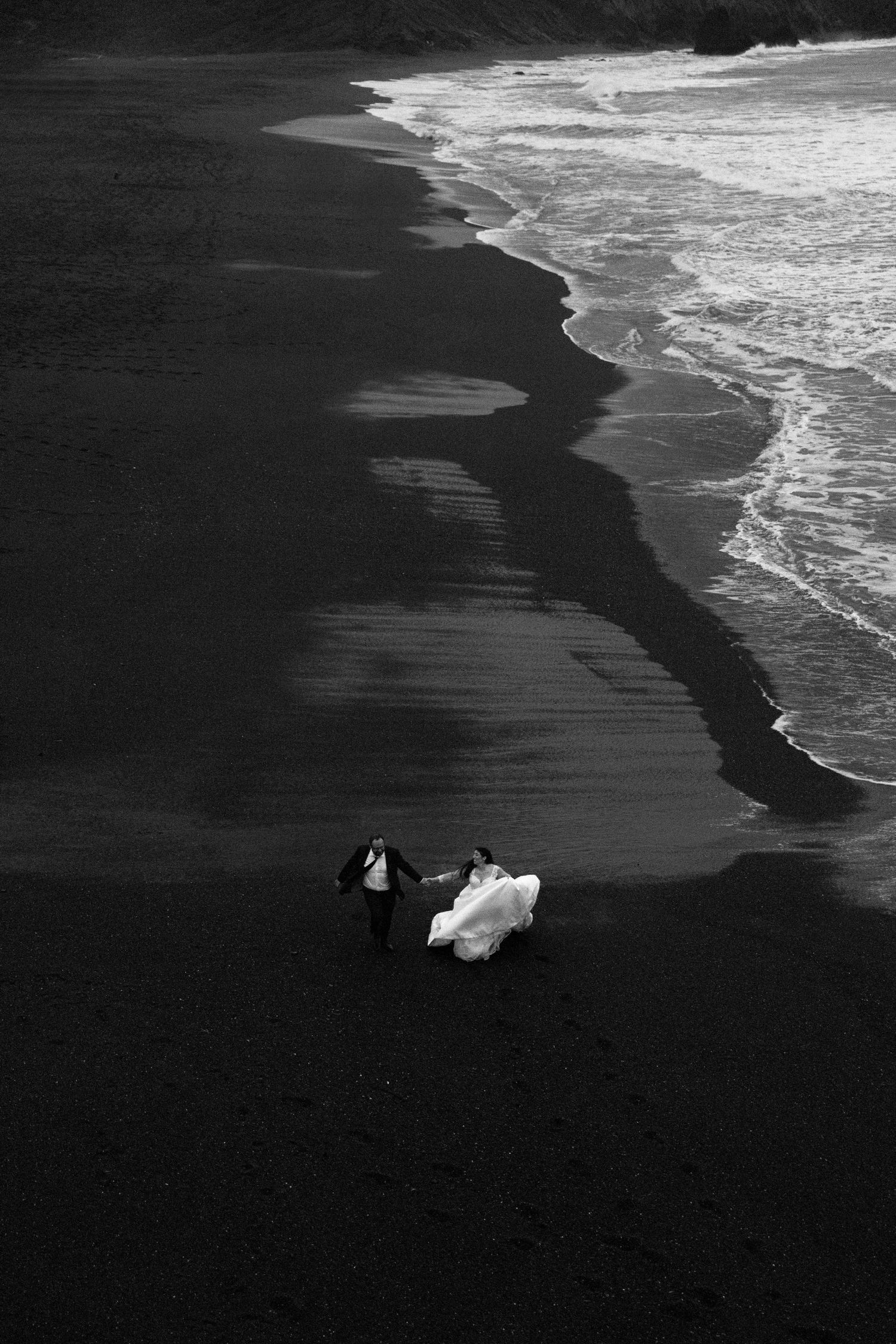 An aerial black and white view of a couple on a dark beach with white waves crashing along the shoreline.