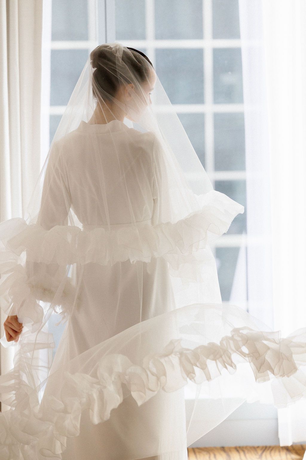 Ethereal bridal portrait with flowing white veil and ruffled dress silhouetted against bright window light.