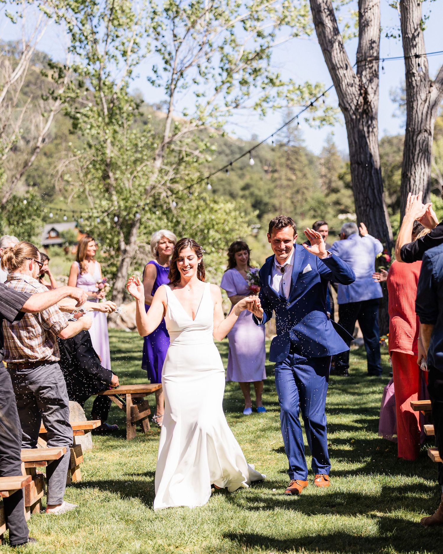 Wedding guests dance and celebrate on a sunny day in a scenic outdoor venue with trees and greenery.