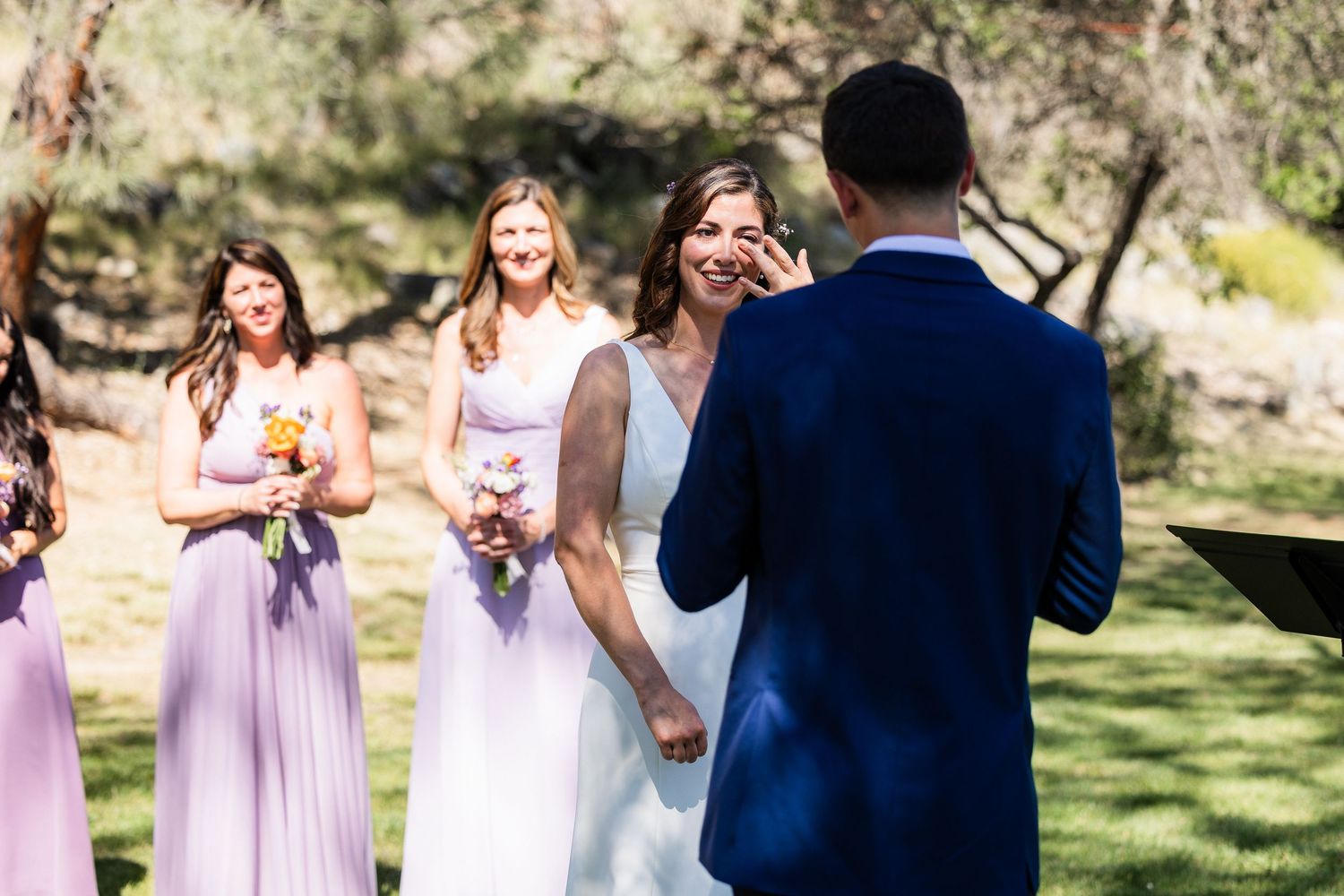 Wedding party in light purple dresses and navy suits gathers outdoors in a sunny garden setting.