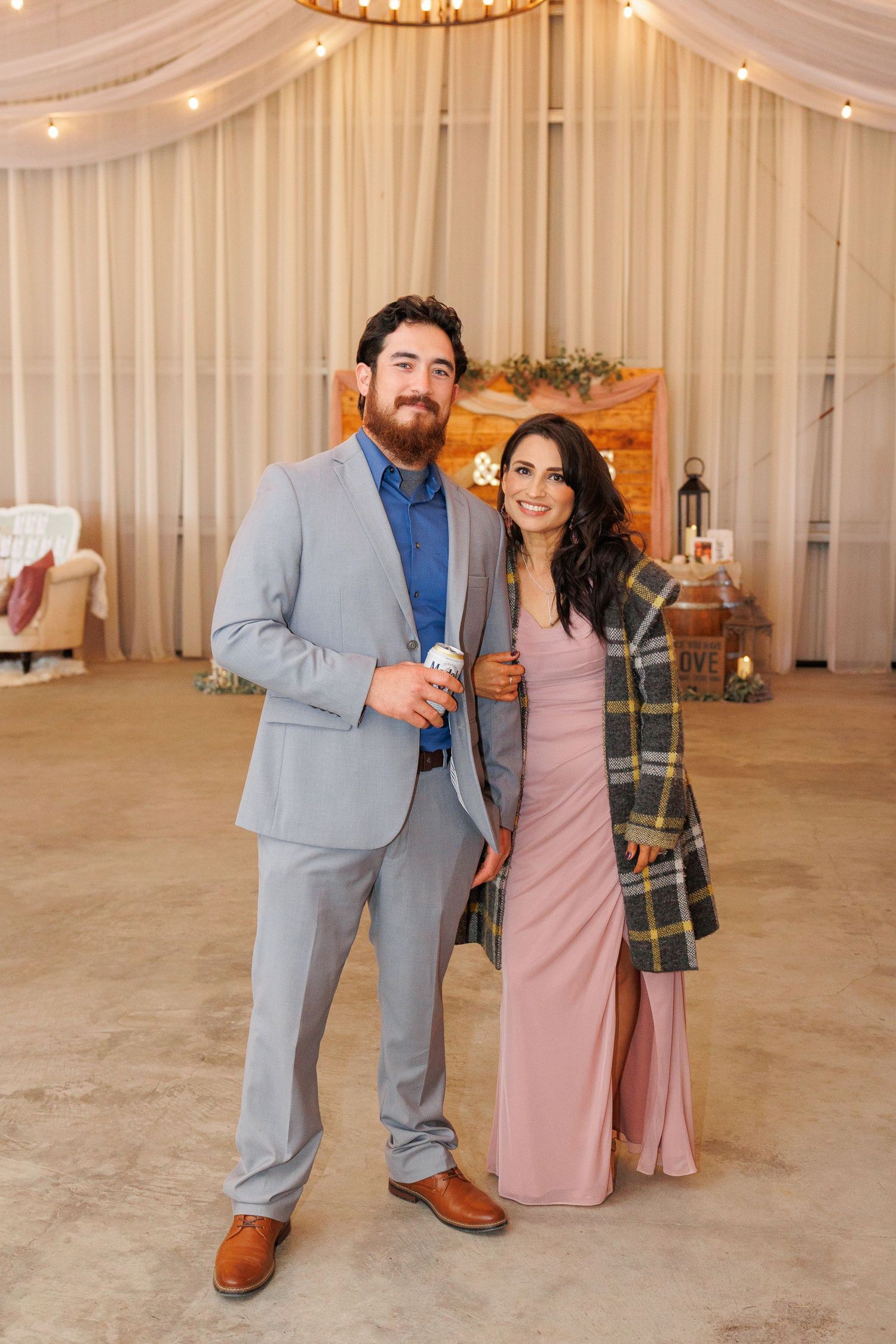 A couple in formal attire poses together in a reception venue decorated with white drapes and chandeliers.