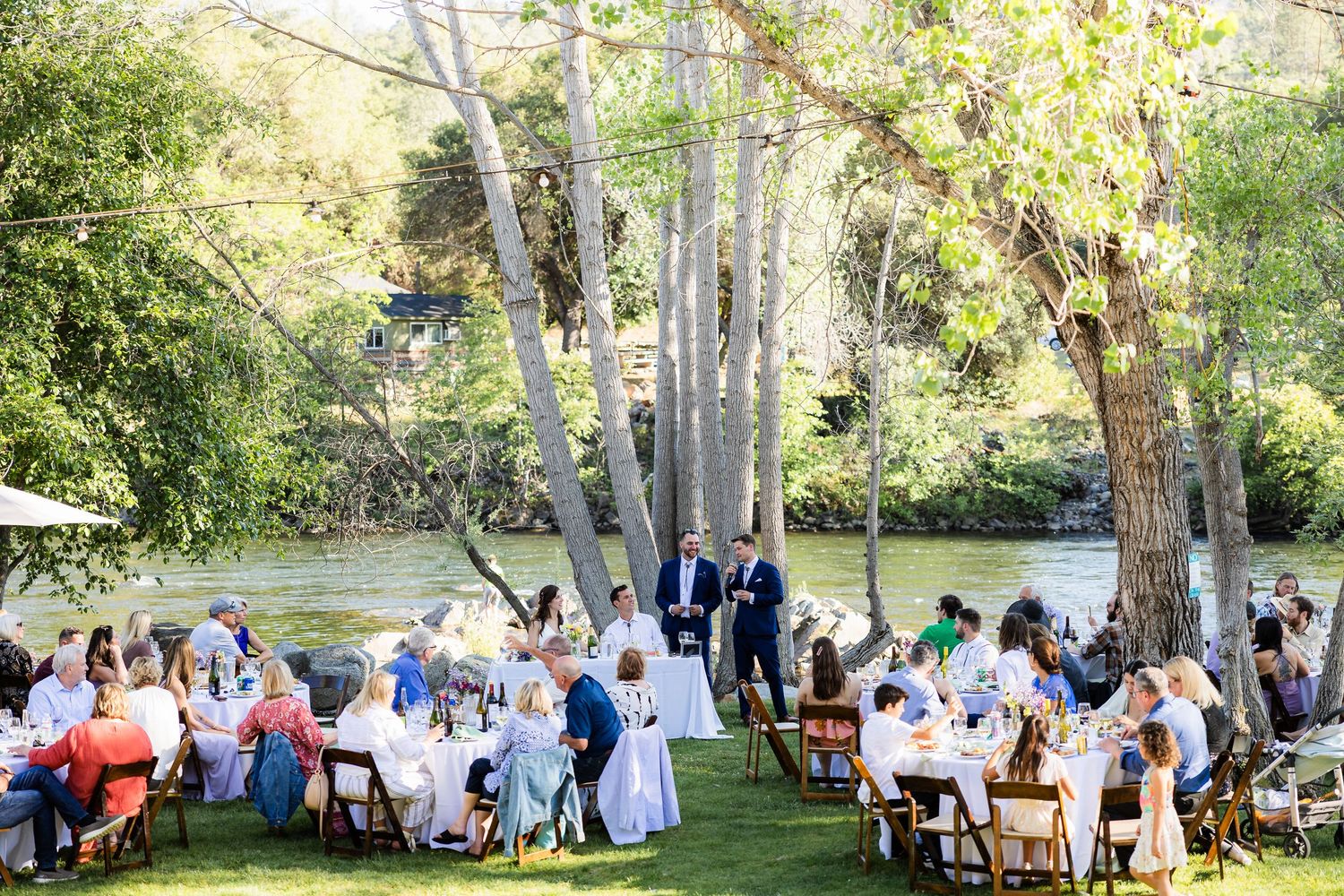Outdoor wedding reception with tables set up under tall trees in a natural garden setting.