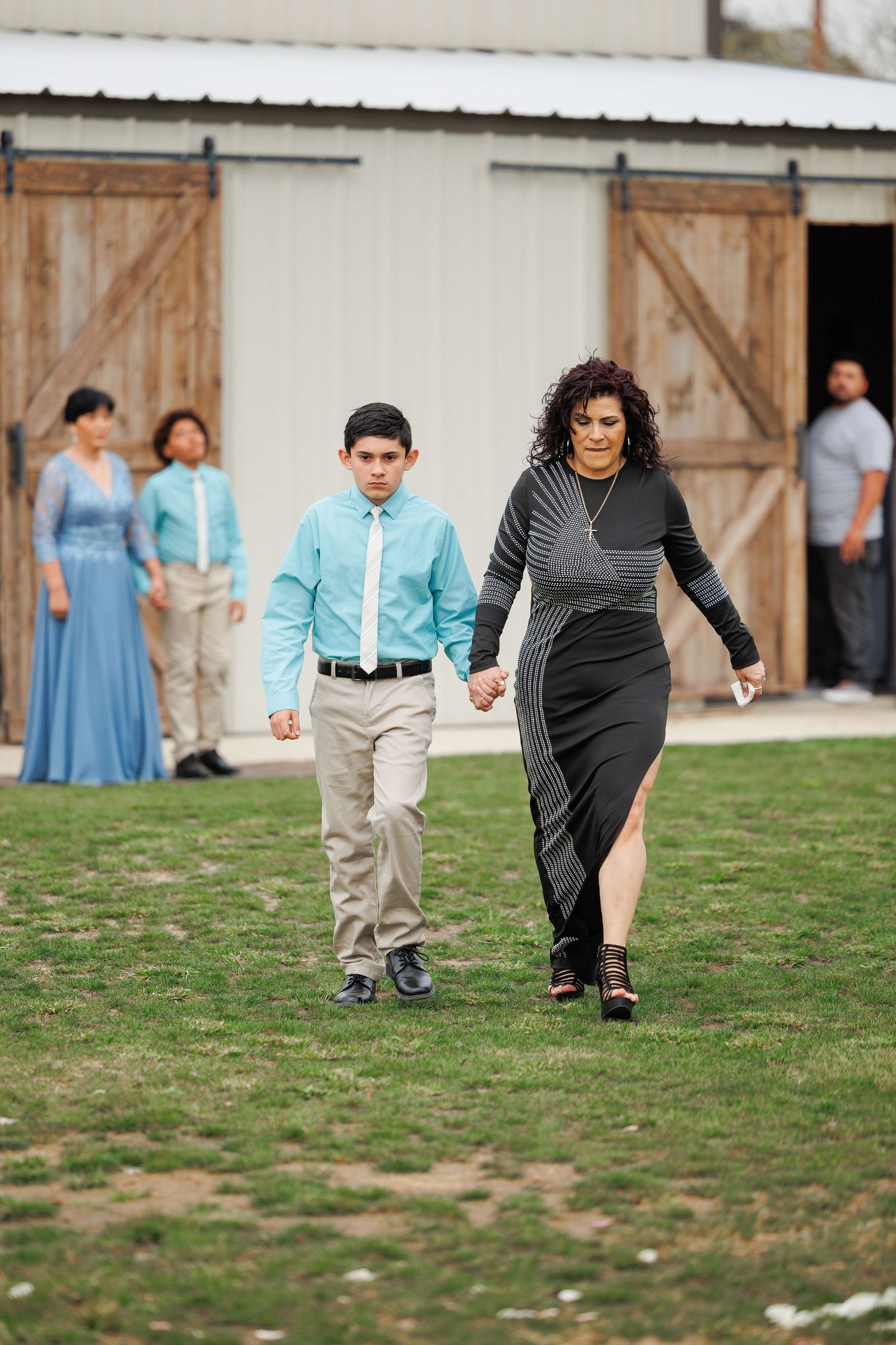 People in formal attire walk across a grassy lawn outside a rustic barn during a special event.