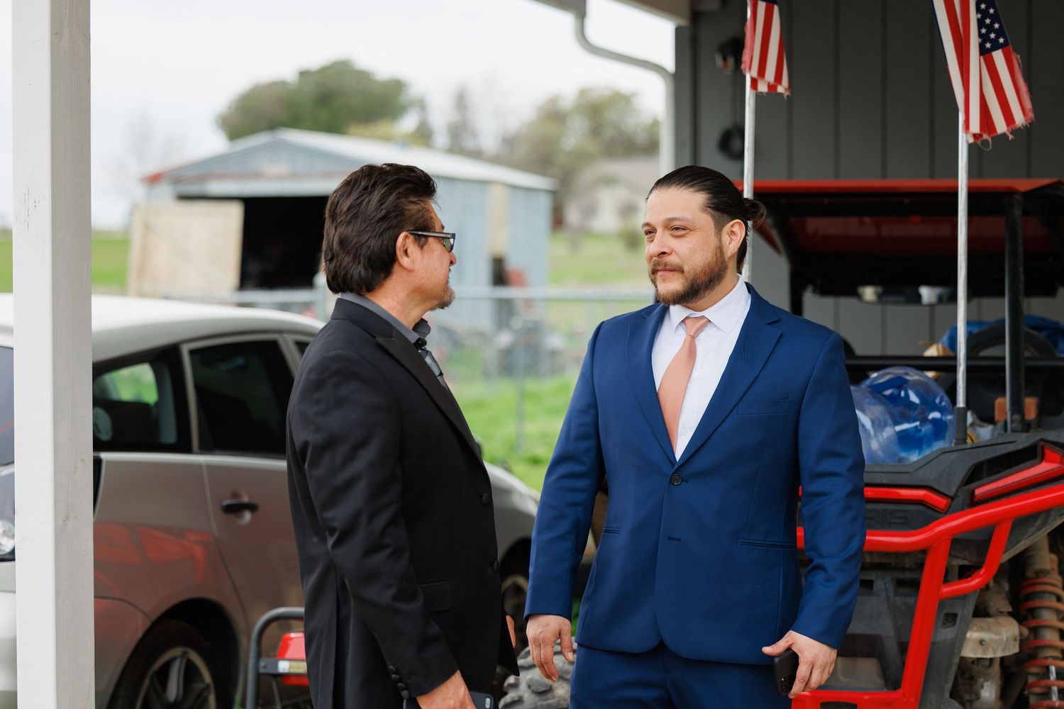 Two people in formal attire stand talking in front of a garage with American flags visible in the background.
