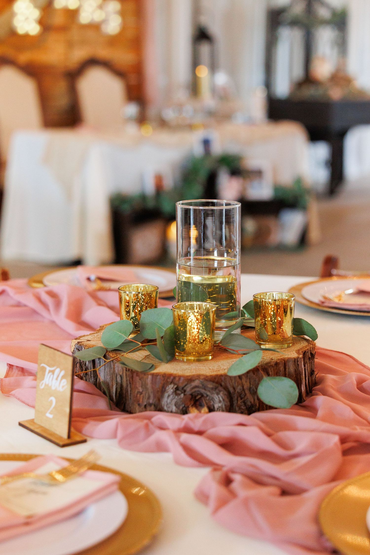 Wedding table decor features rustic wood slices, gold votives, and pink tablecloths with eucalyptus accents.