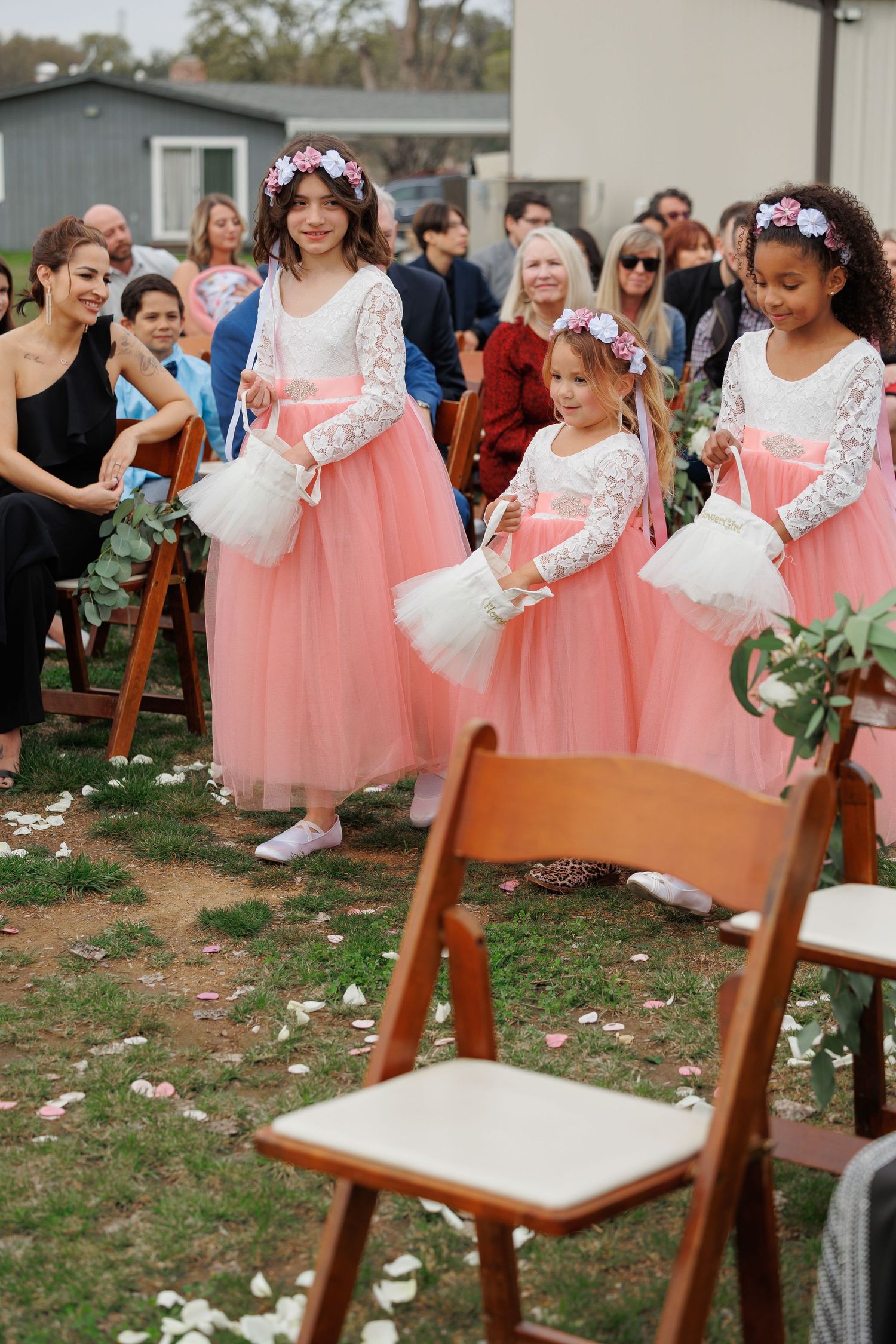 Flower girls in matching pink tulle dresses and white lace tops carry baskets at an outdoor wedding ceremony.