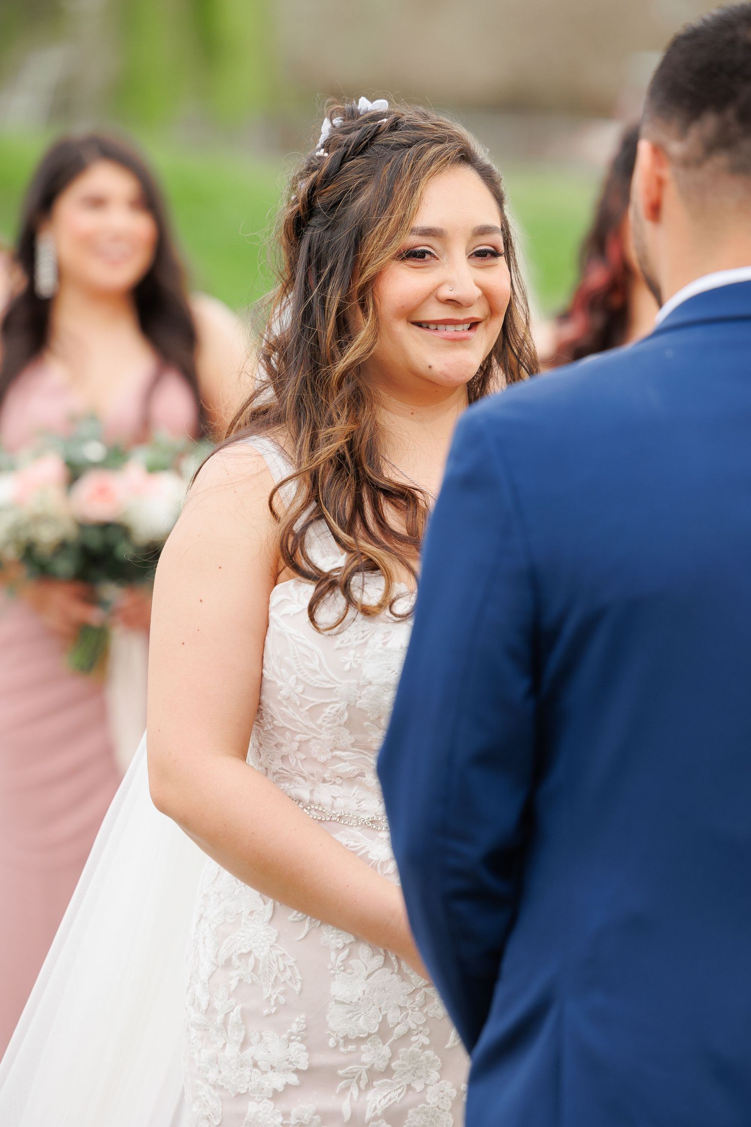 Bride in white lace wedding dress smiles joyfully during outdoor wedding ceremony with guests in background.