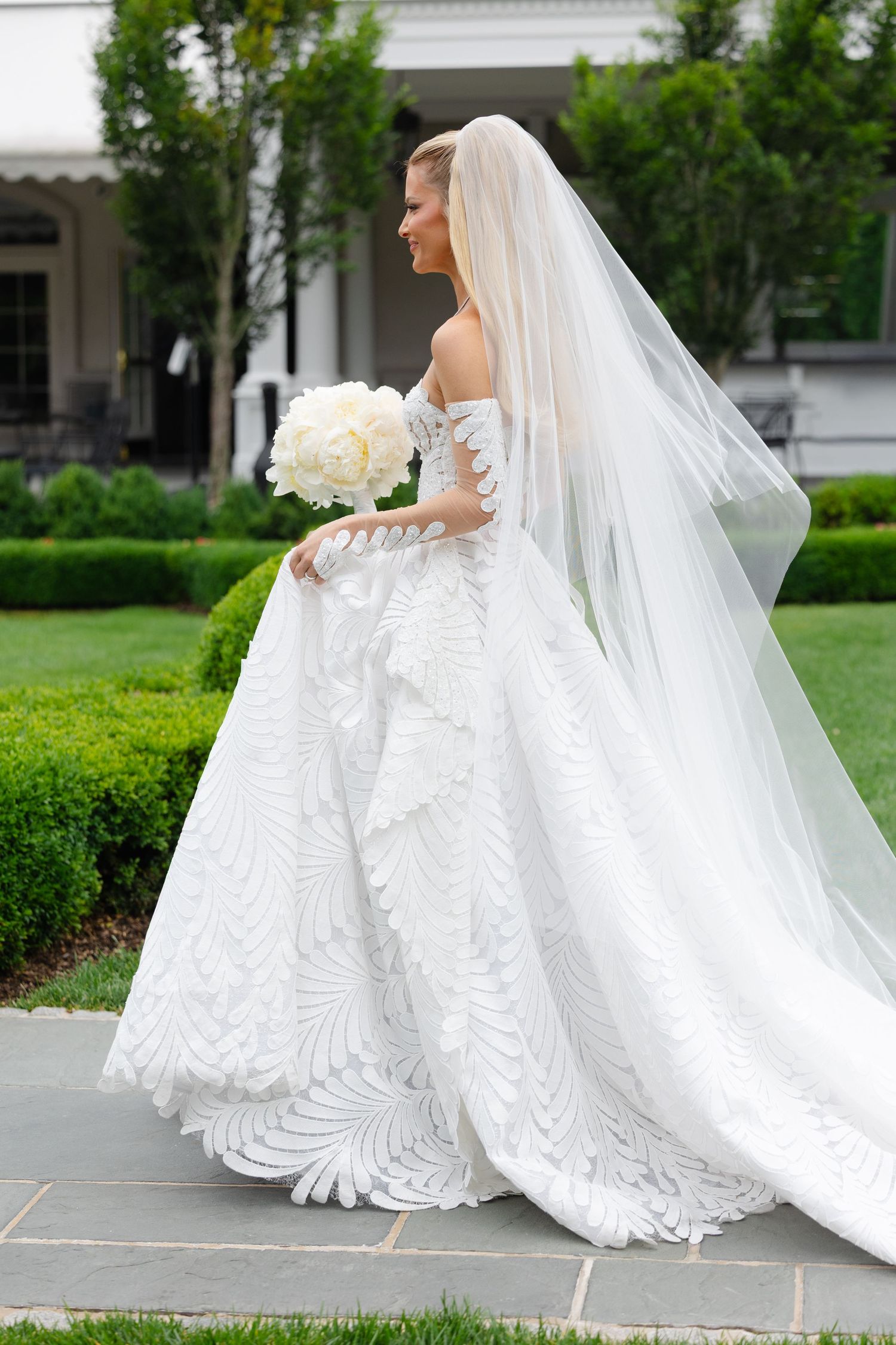 A bride in an elegant white lace wedding gown with a flowing veil poses gracefully in a garden setting.