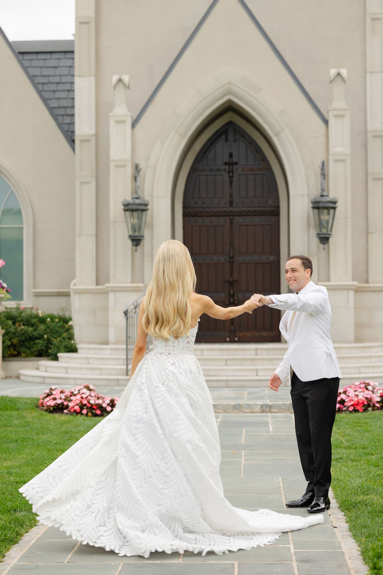 A couple in wedding attire dances together outside a Gothic-style church with blooming pink flowers.