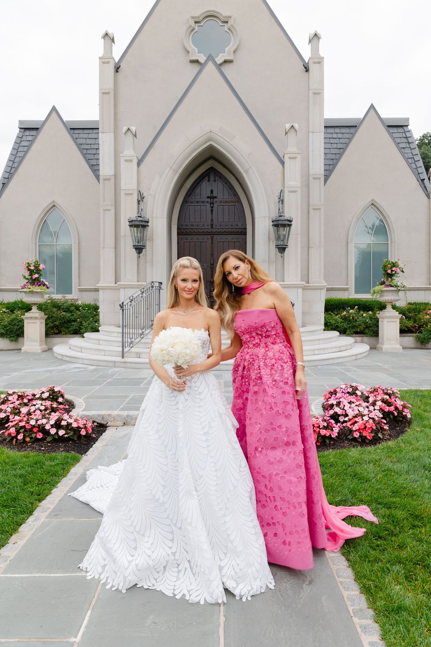 Two women in formal dresses pose together outside a Gothic-style church with white stone walls and pink flowers.