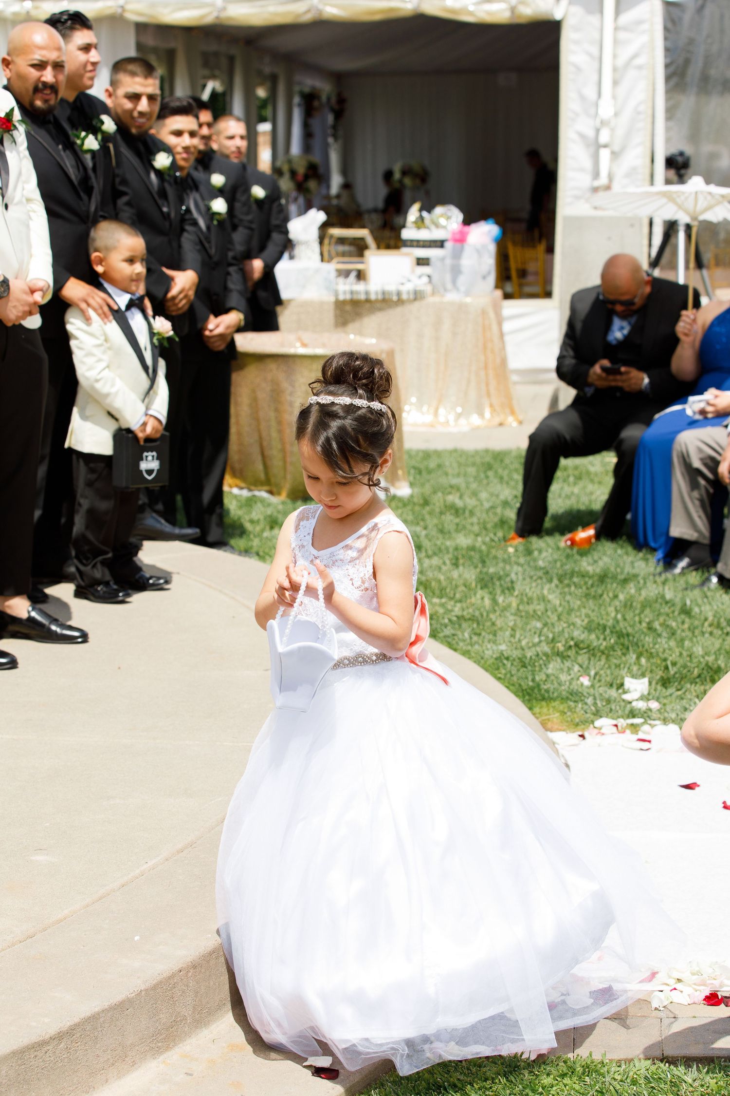 A child in a white dress sits on a lawn during an outdoor event while guests mingle in the background.