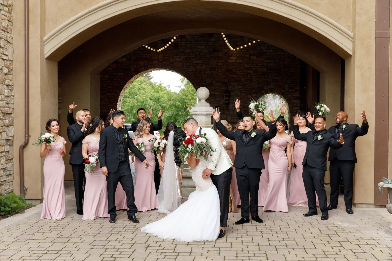 Wedding party in formal attire poses under stone archway with bridesmaids in pink dresses and groomsmen in black suits.