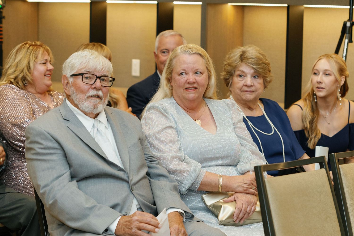 Audience members seated in a row at a formal event with gold-toned decor.