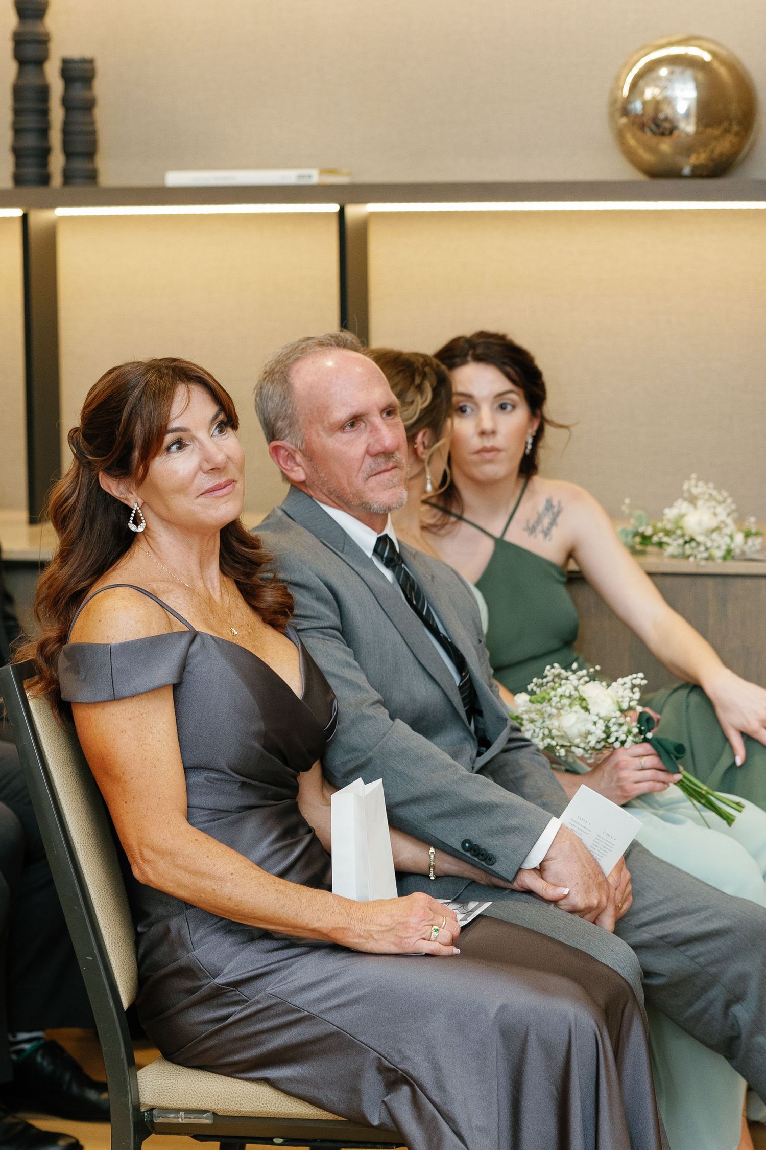 Wedding guests in formal attire sit together, with women wearing gray and green dresses and a man in a gray suit.