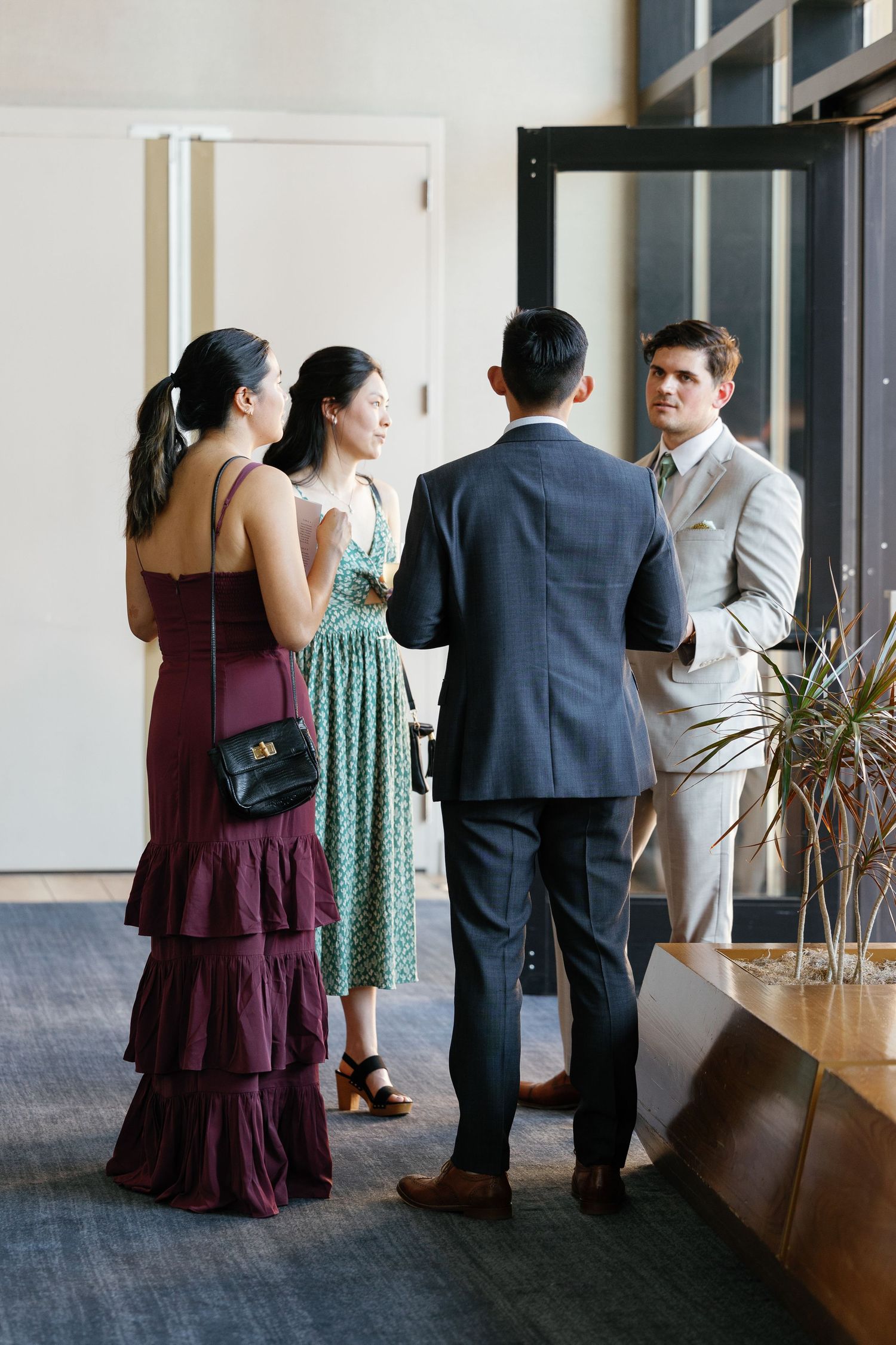 People in formal attire gather and mingle at an indoor event space with large windows and carpeted floors.