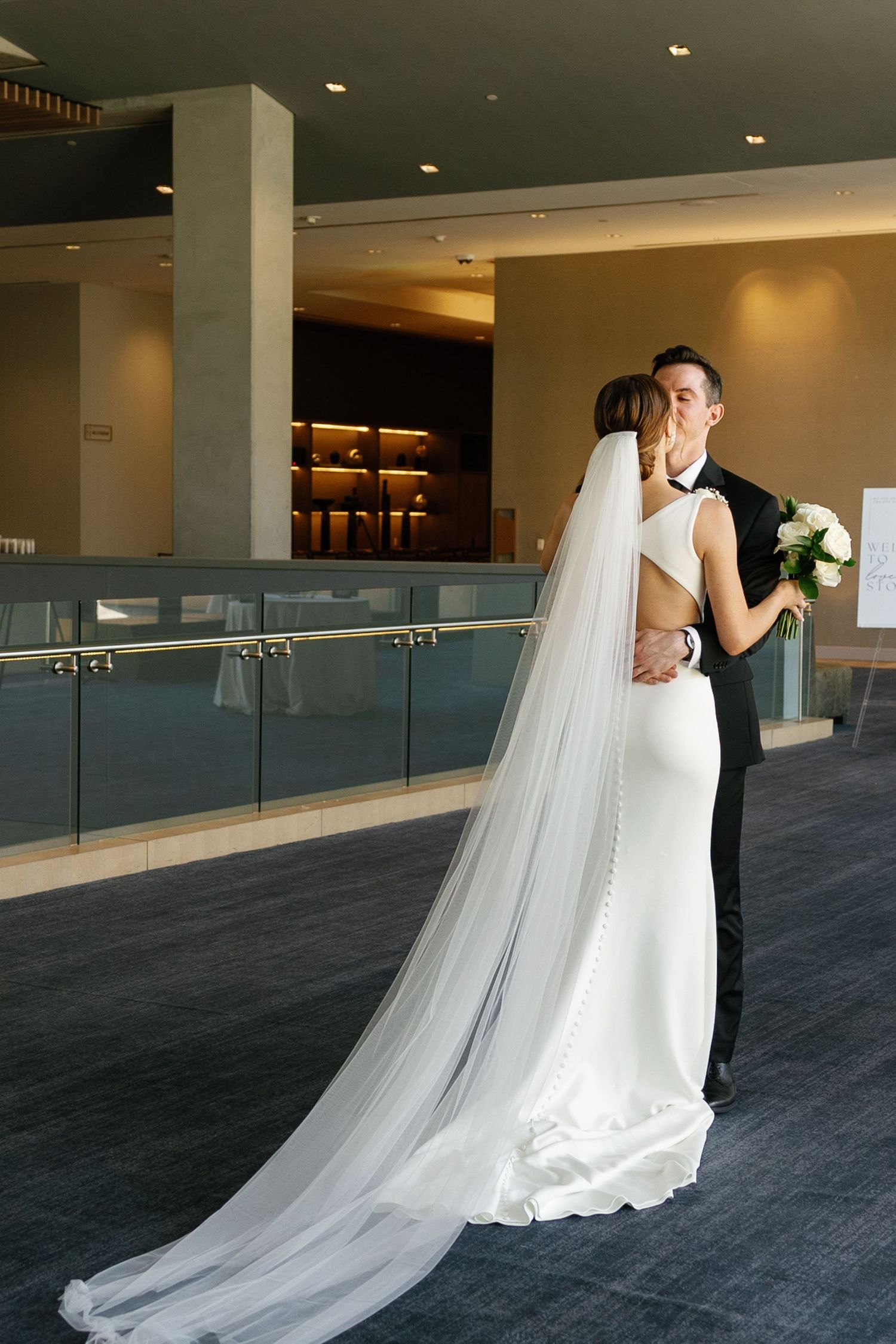 A bride in a flowing white wedding gown with a long cathedral veil stands in an elegant modern hallway.