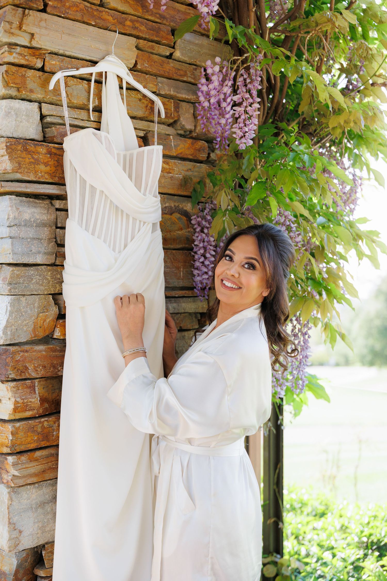 A white wedding dress hangs elegantly against a stone wall with blooming wisteria flowers cascading above.