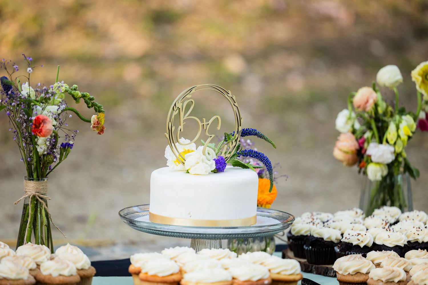White frosted wedding cake decorated with fresh flowers and greenery on a rustic wooden table.