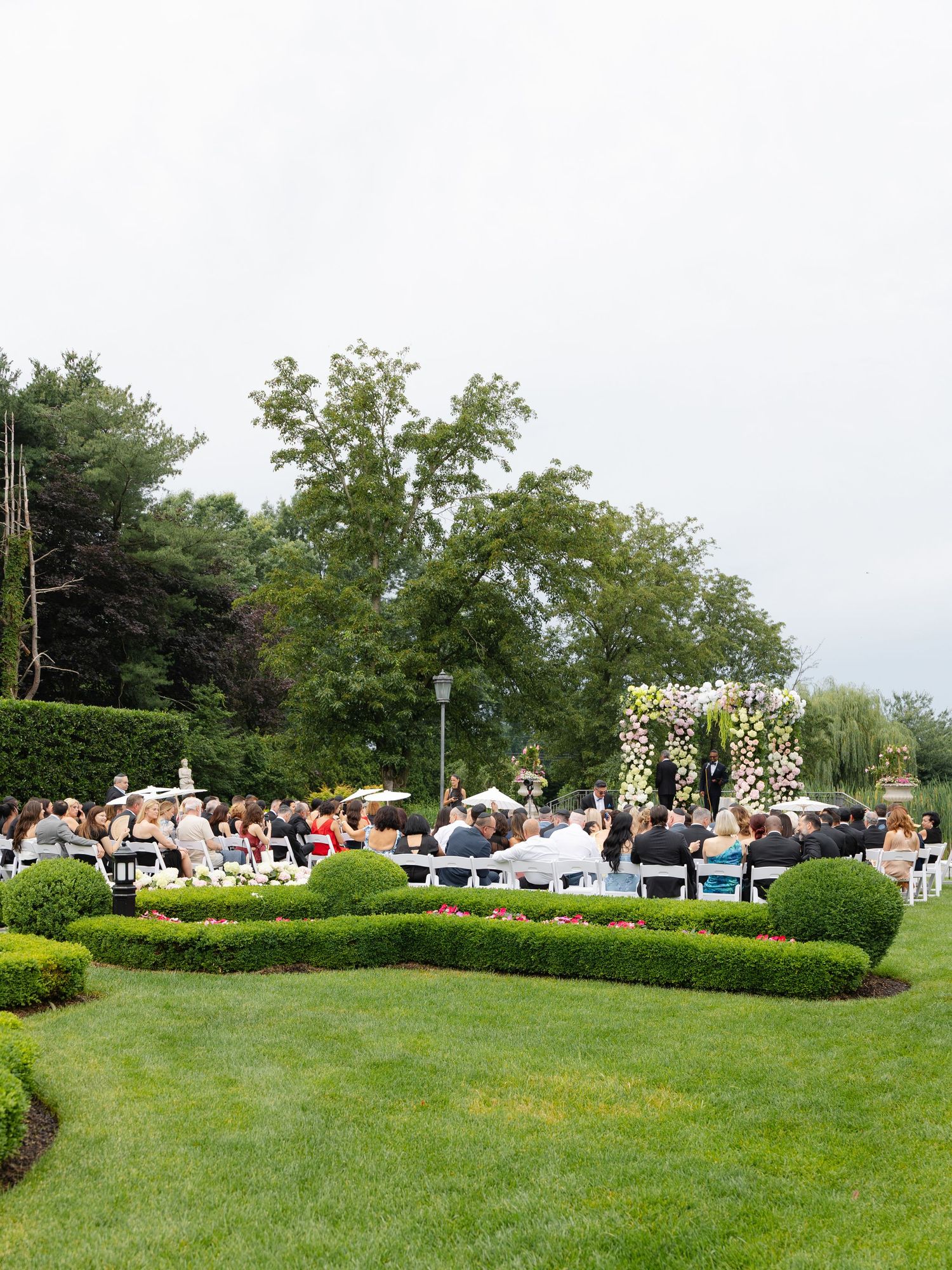 Elegant outdoor wedding ceremony in lush garden with floral arch, white chairs, and guests seated on manicured green lawn.