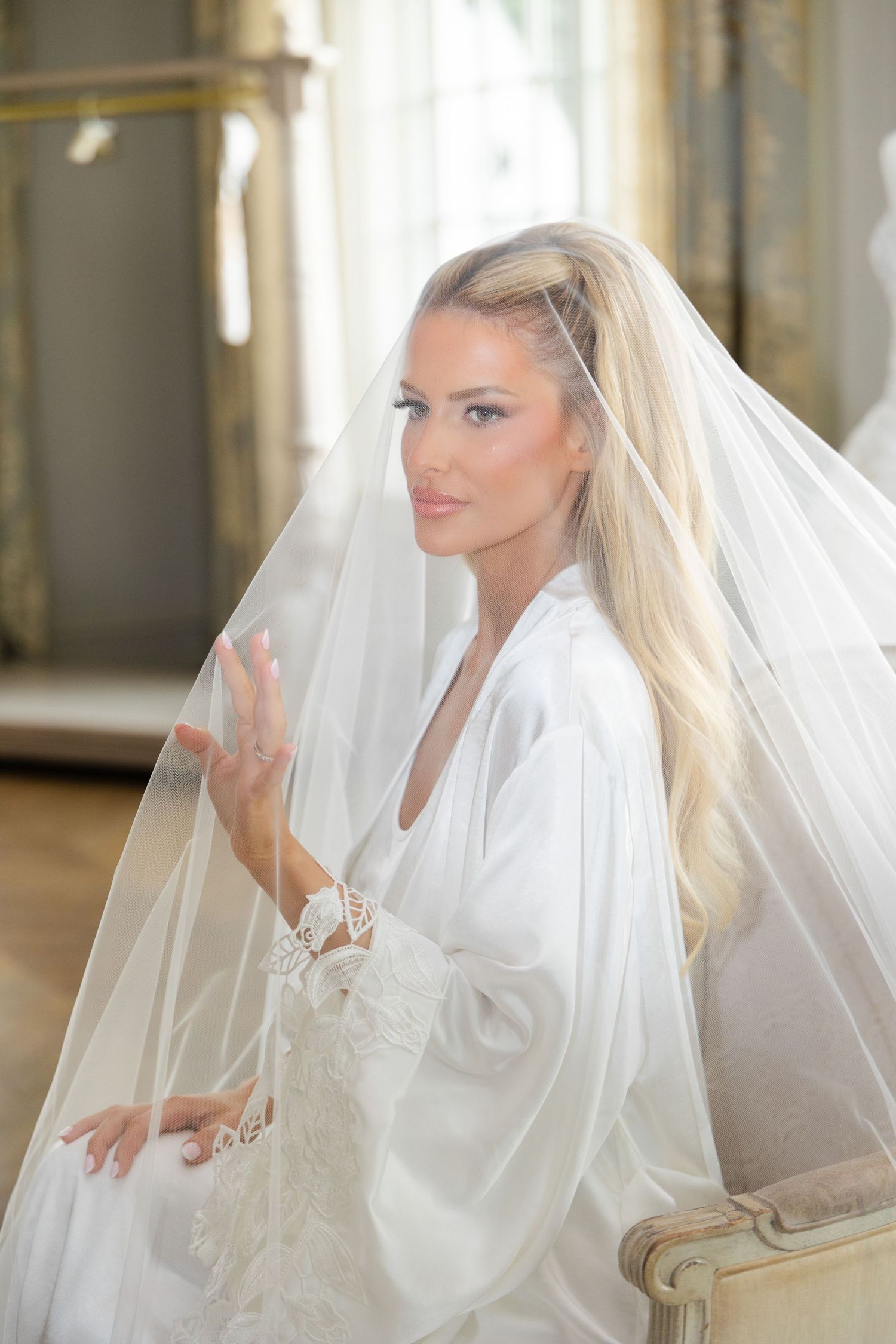 Blonde bride in white robe and veil sitting elegantly in a chair during bridal preparations.