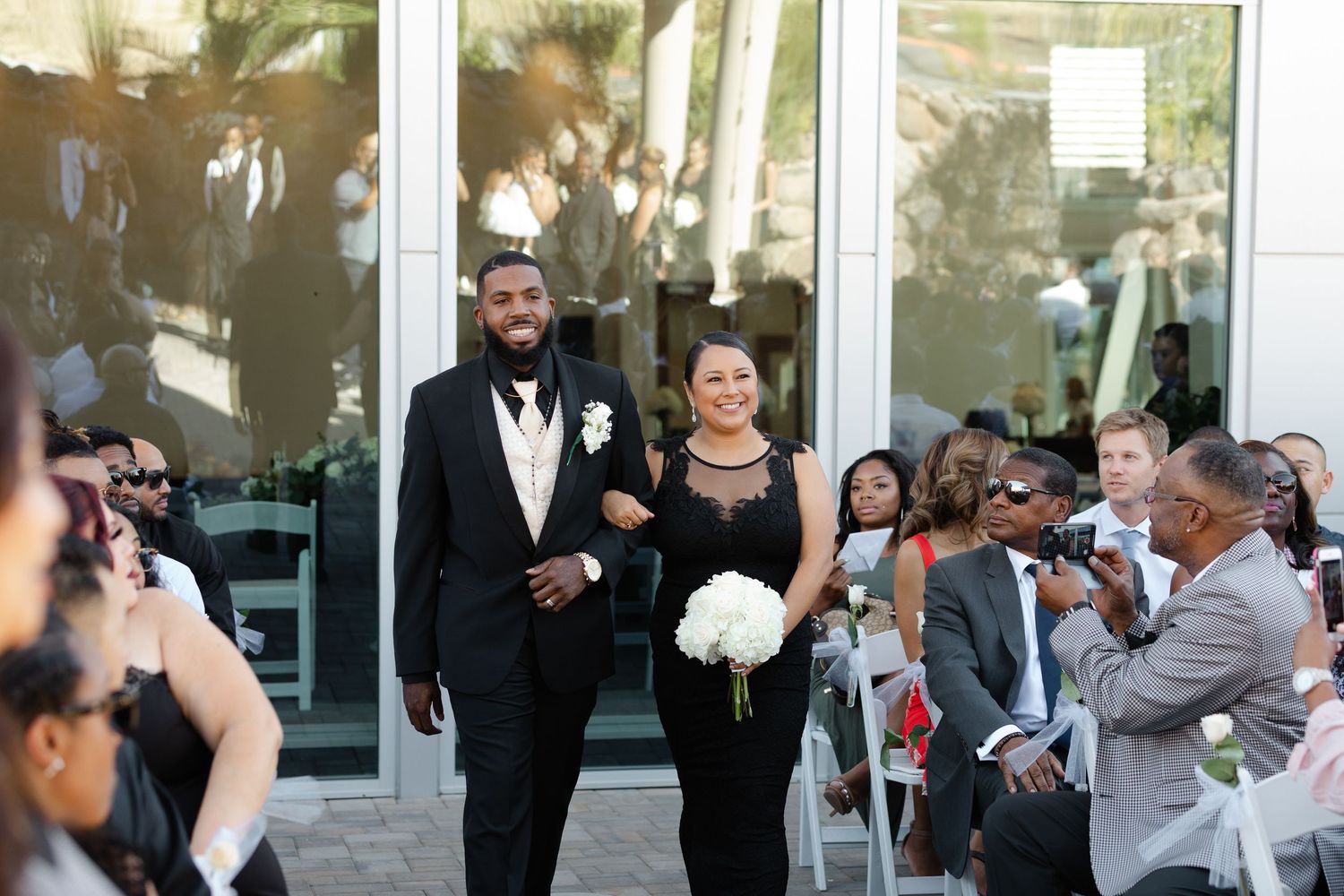 Wedding party members in formal attire exit a glass door building while guests look on during an outdoor ceremony.