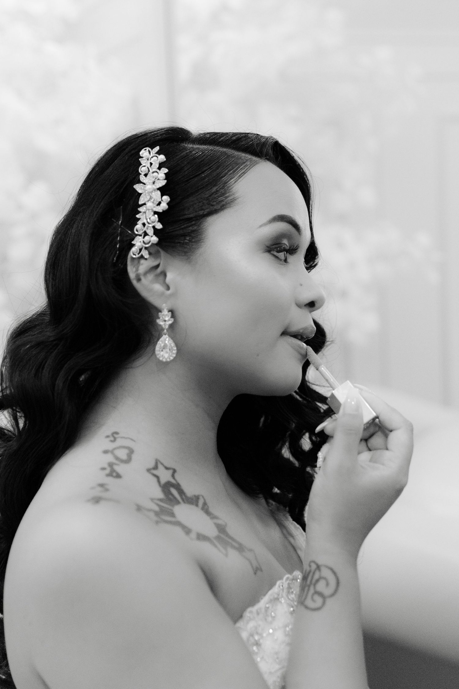 Black and white portrait of a bride applying lipstick, wearing a floral hair accessory and crystal drop earrings.